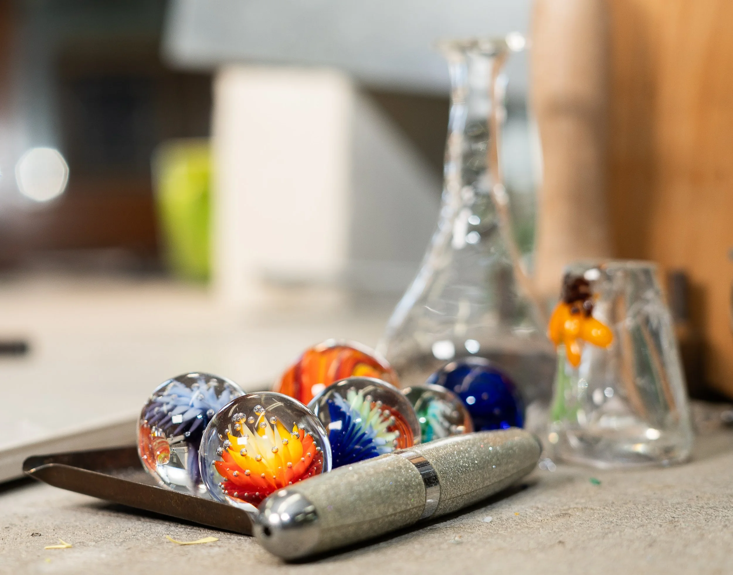 Close-up of a metal tray with colorful glass marbles, in front of glass pipes on a countertop.