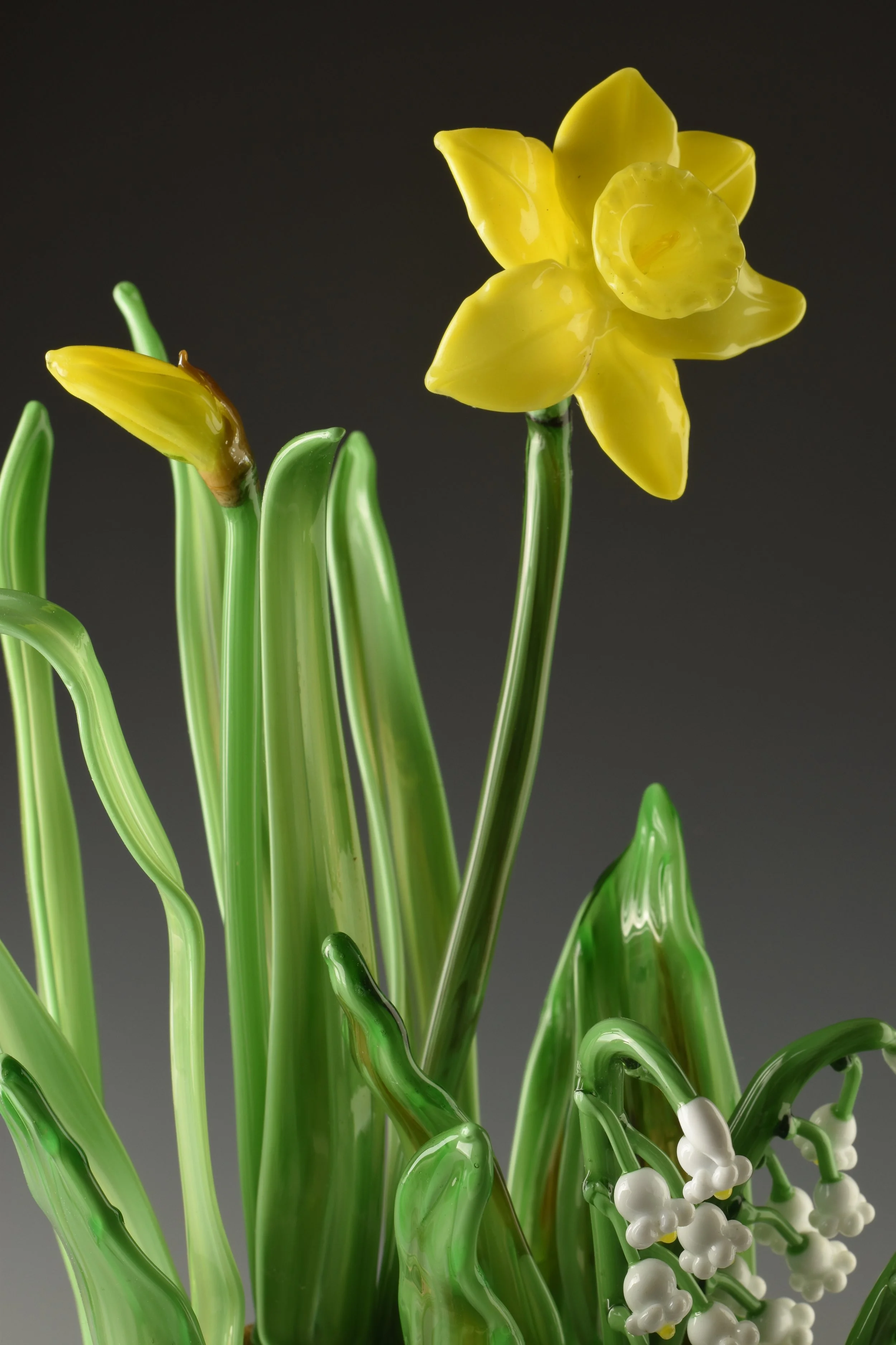 Glass sculpture of yellow daffodil flowers with green stems and white accents on a dark background.