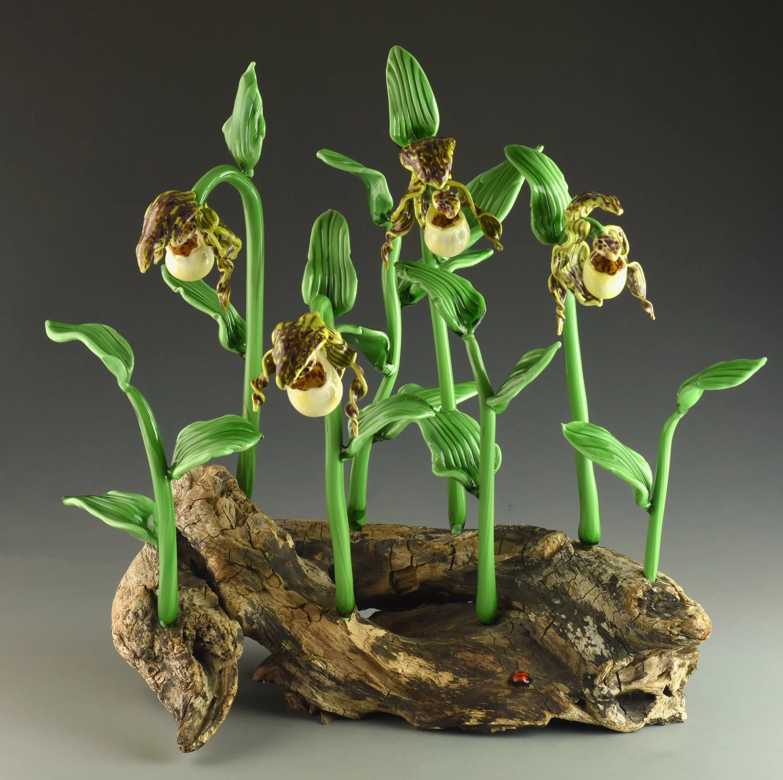 A sculpture of orchids with green stems and leaves, yellow and brown patterned flowers, and a small ladybug on a piece of wood at the bottom.