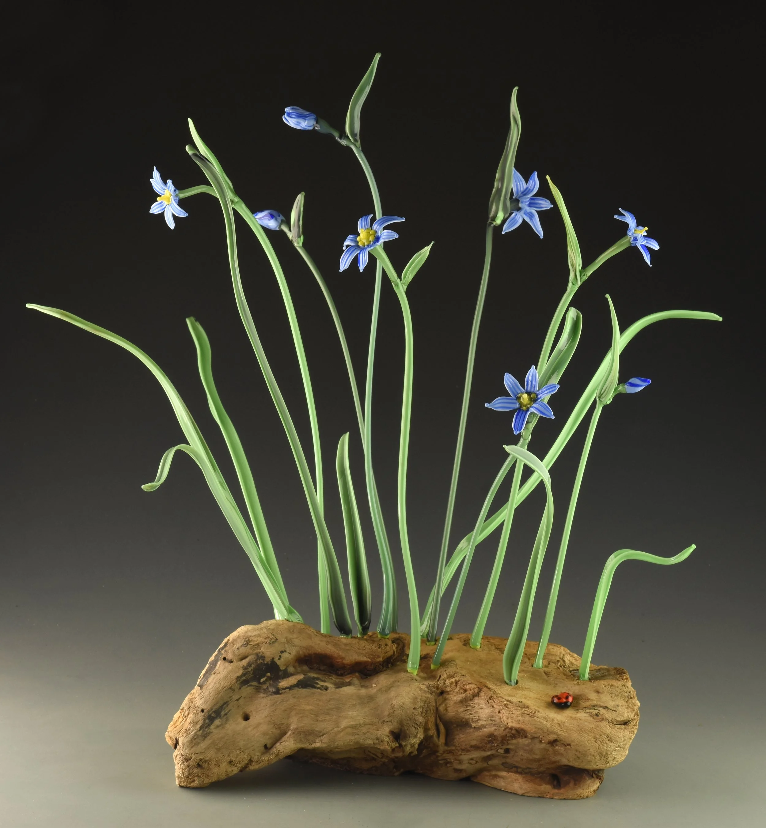 Glass sculpture of blue flowers with green stems on a textured wooden base, featuring a tiny ladybug at the bottom right.