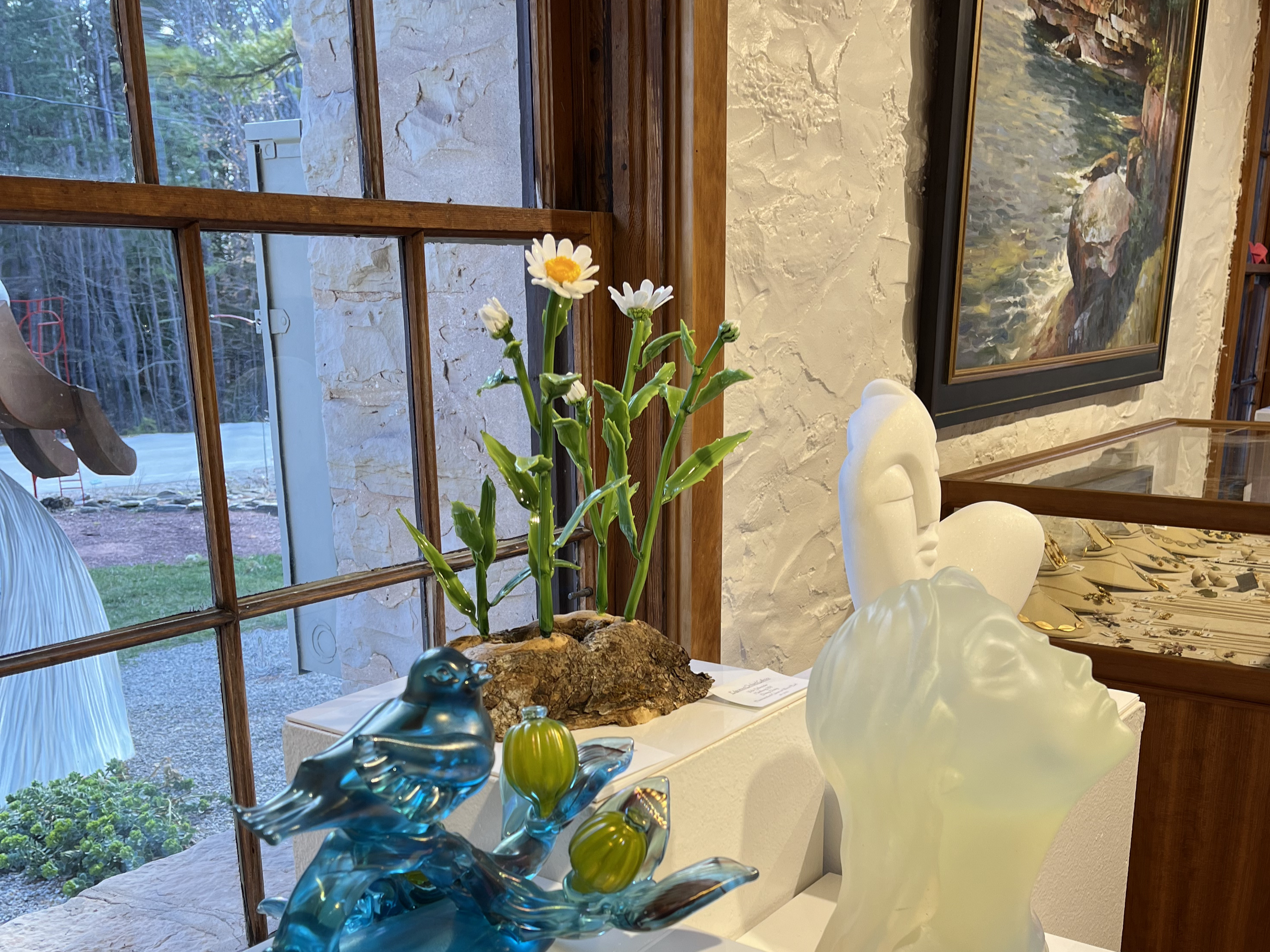 Interior of a room with stone walls, wooden window frames, and a landscape painting hanging on the wall. A glass sculpture of a bird and a decorative vase are on the surface, with a small bouquet of daisies in a pot near the window. Outside the window, trees and a yard are visible.