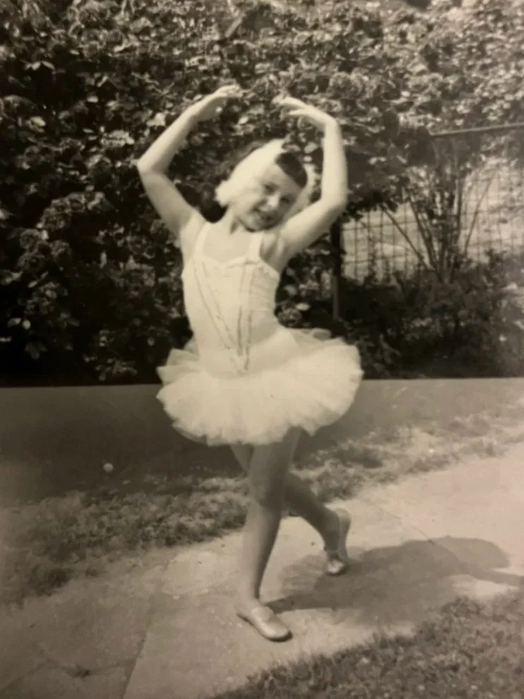 A young girl in a ballet tutu raises her arms above her head, standing on one foot outdoors near bushes on a sunny day.