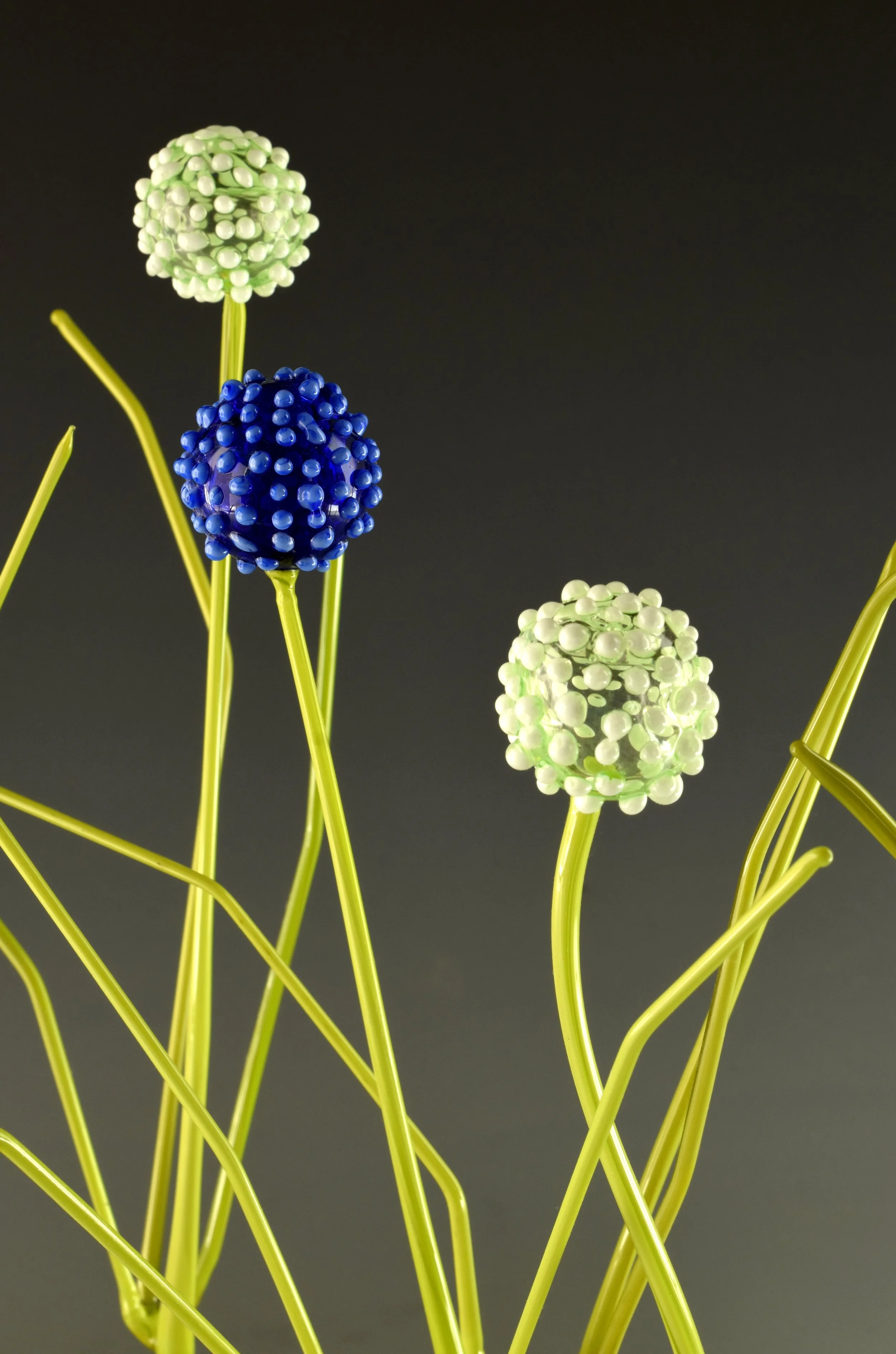 Artificial flowers resembling alliums with green and blue spherical buds on yellow stems against a dark background.