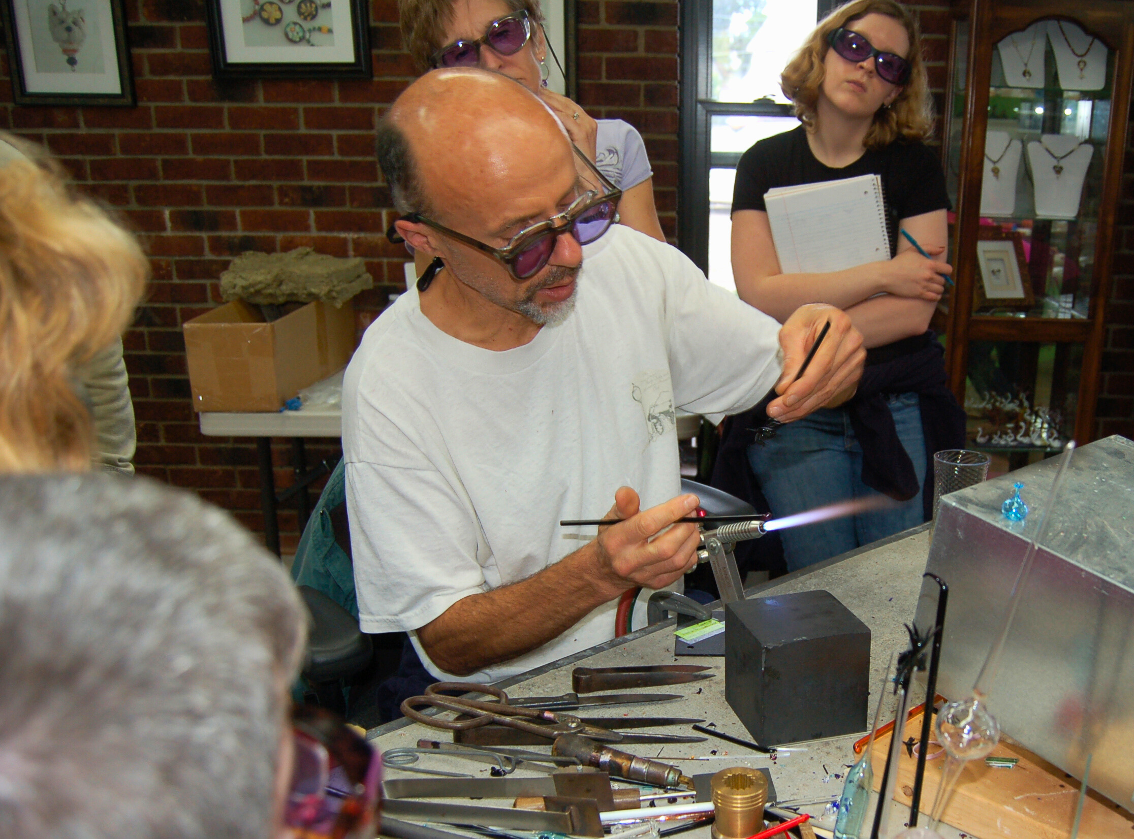 A man with a bald head and glasses, wearing a white shirt, is working with tools at a craft or jewelry-making station. Two women are standing behind him, observing him work. The woman on the left has short hair, glasses, and is wearing earrings, while the woman on the right has curly hair, sunglasses, and is holding a notebook. The workspace is set against a brick wall decorated with framed artwork, and various tools and materials are on the table.