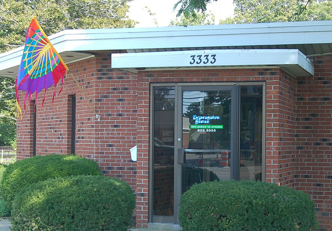 Brick building with a glass door, green bush plants in front, and a colorful flag on the left side. The building has the number 3333 above the door and a neon sign on the door that reads "Expressive glass, the place to create, 826.3333."