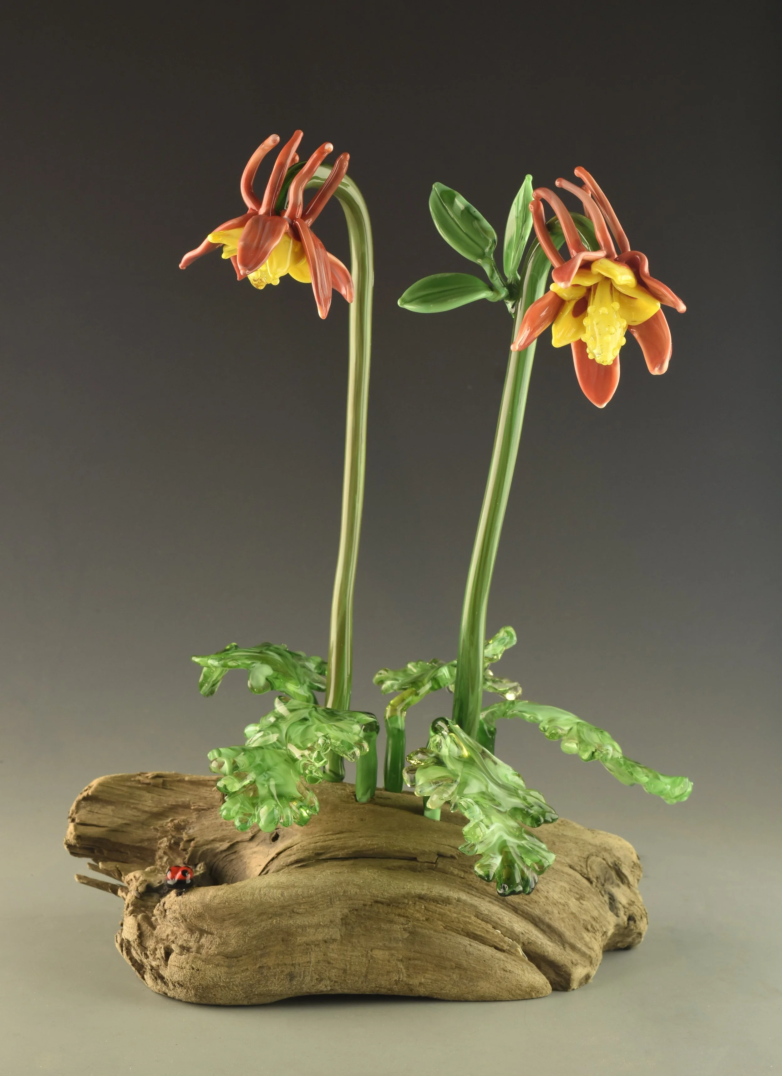 Glass sculpture of a flowering plant with yellow and orange flowers on a piece of wood with a small ladybug at the bottom.