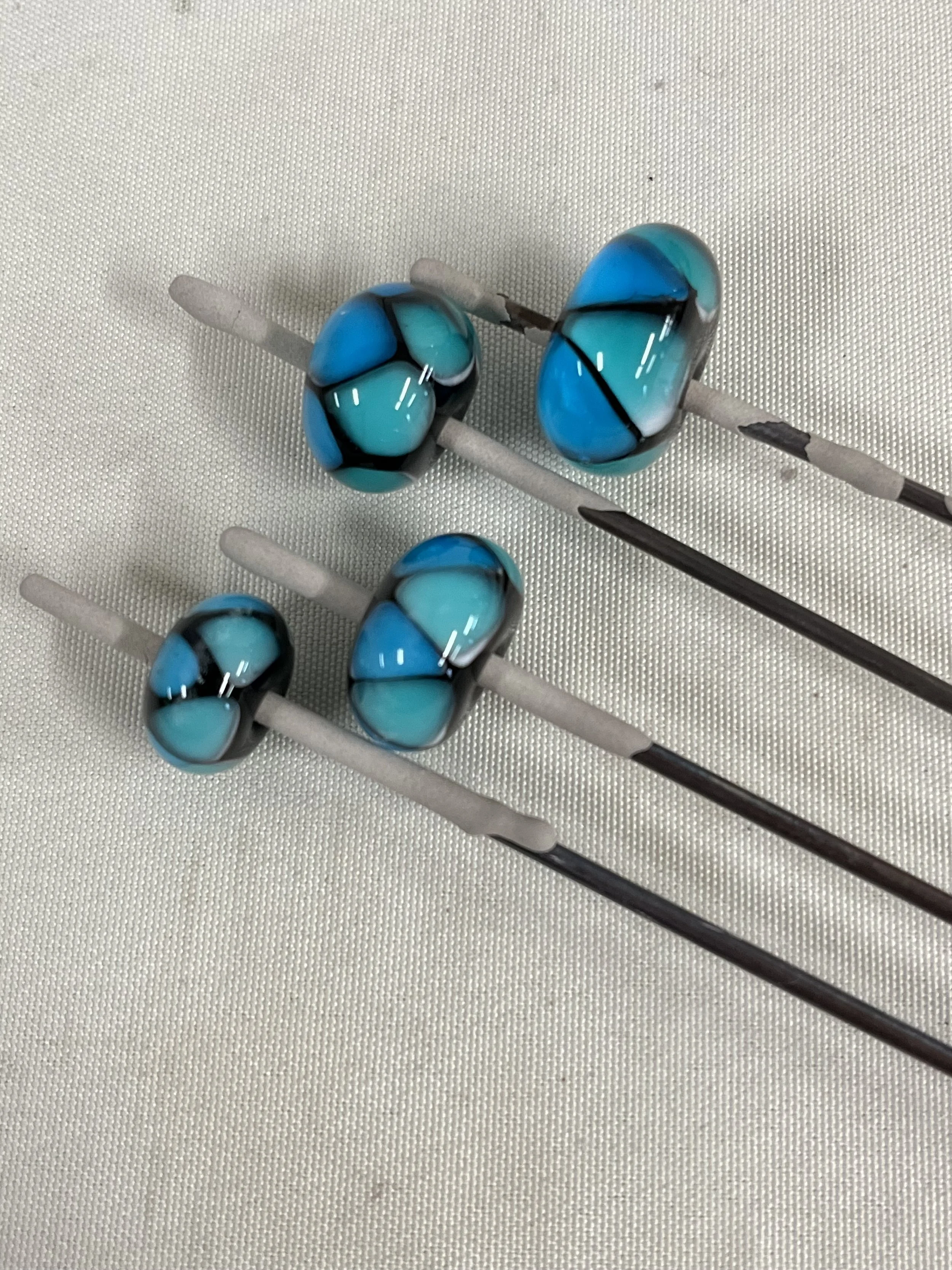 Close-up of glass beads with black and white swirls on thin metal sticks, resting on a textured white surface.