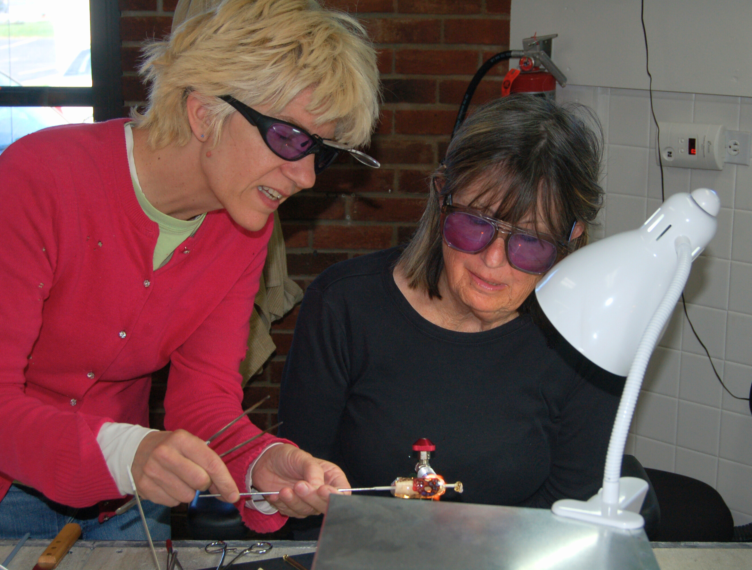 Two women, wearing protective glasses, are working together on a scientific or technological experiment involving a small robotic device with wires and lights, in a laboratory setting.