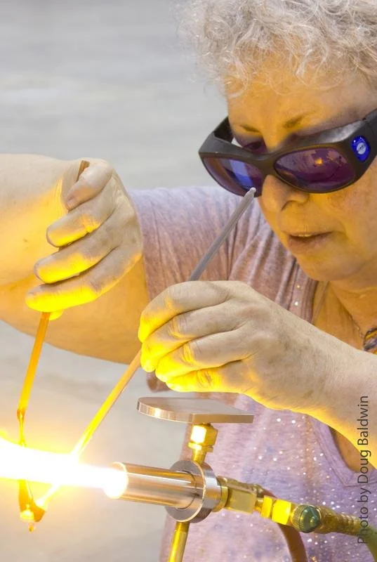 An elderly woman with curly gray hair and sunglasses welds metal using a welding torch, with bright sparks and yellow safety gloves.