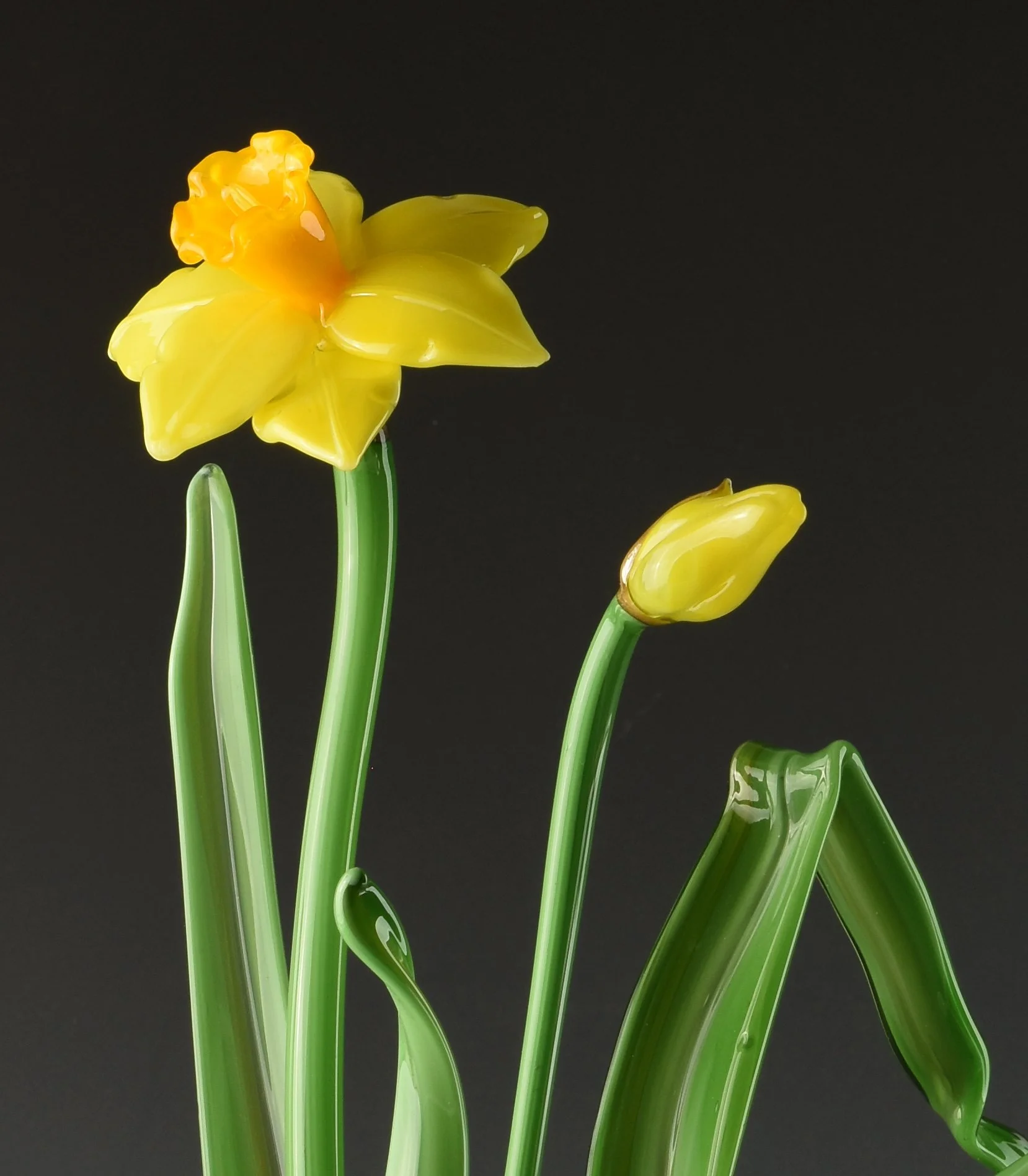 A glass sculpture of yellow and orange daffodil flowers with green stems and leaves against a black background.