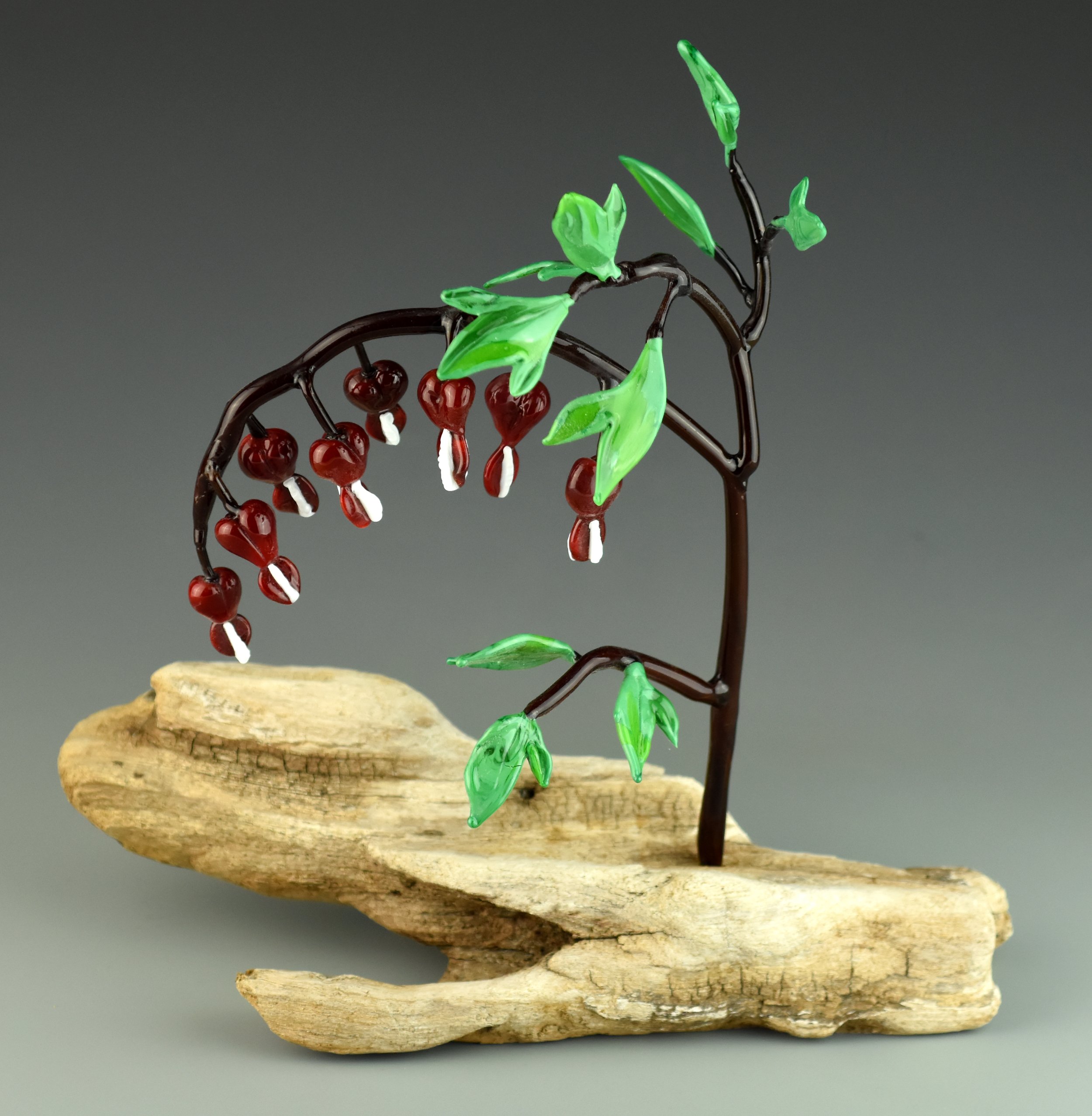 A decorative sculpture of a bird perched on a branch made of twisted dark metal, with green glass leaves and red glass berries, mounted on a natural wood base.