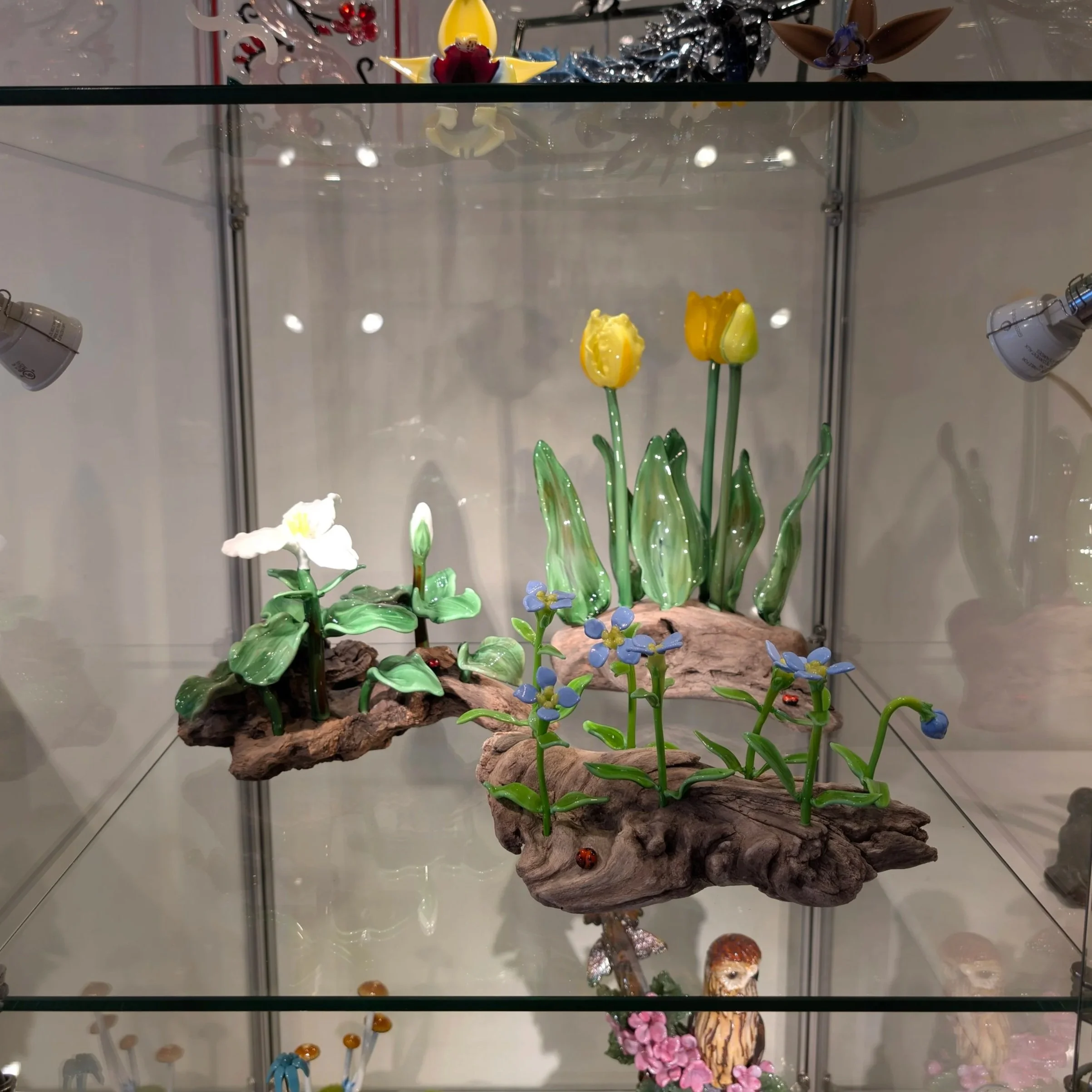 Glass display case with colorful glass art sculptures of flowers and animals, including tulips, roses, and butterflies, on a wooden base, inside a well-lit room.