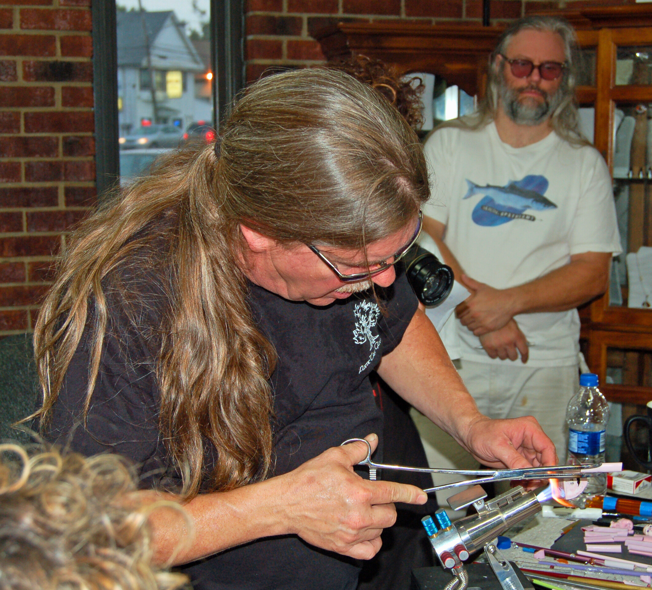 A person with long, wavy hair and glasses working with a tool at a table, with another person in the background wearing glasses and a graphic t-shirt, inside a room with brick walls.