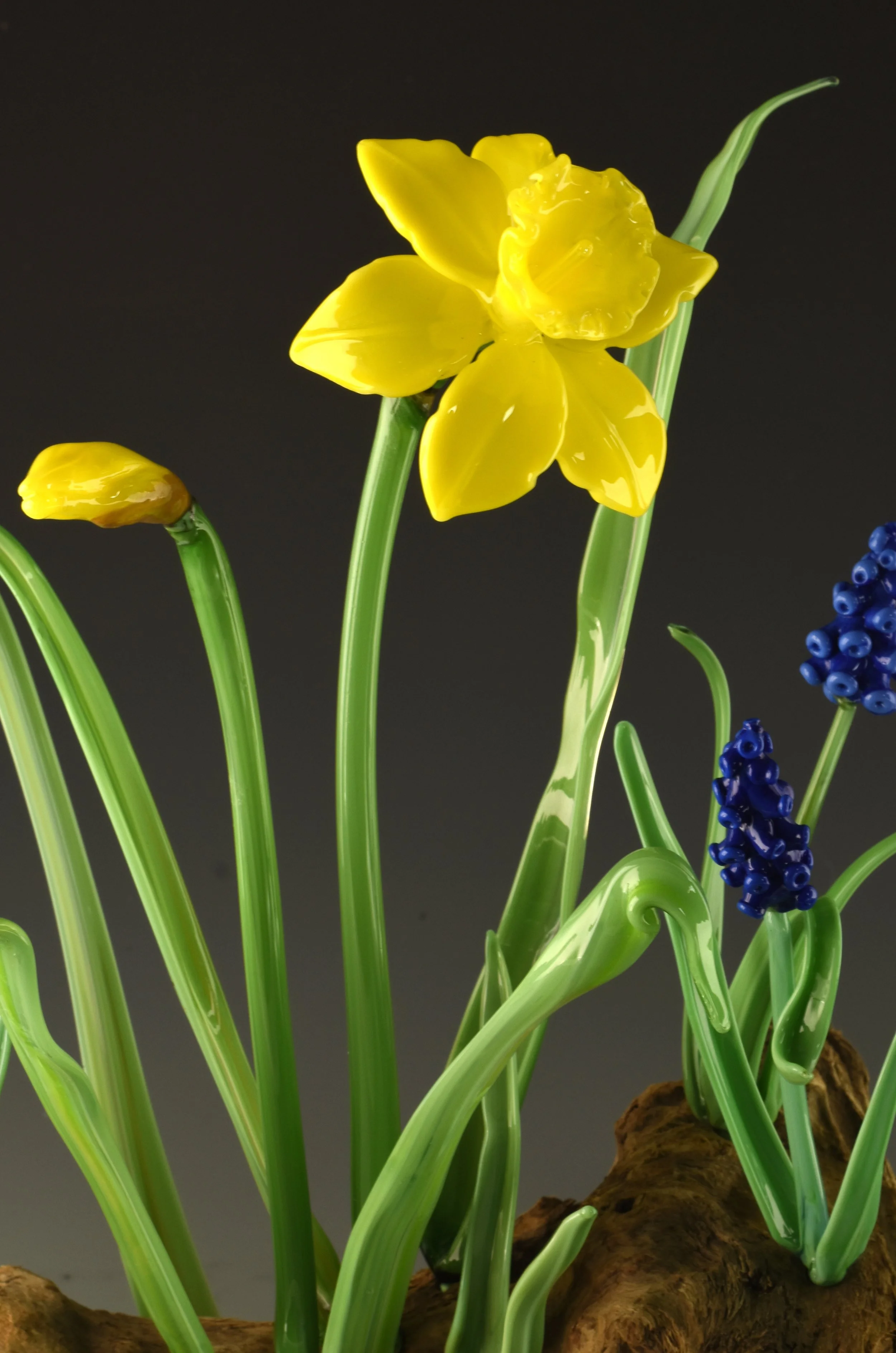 Glass sculpture of yellow flowers, green stems, and blue buds on a rocky base against a dark background.