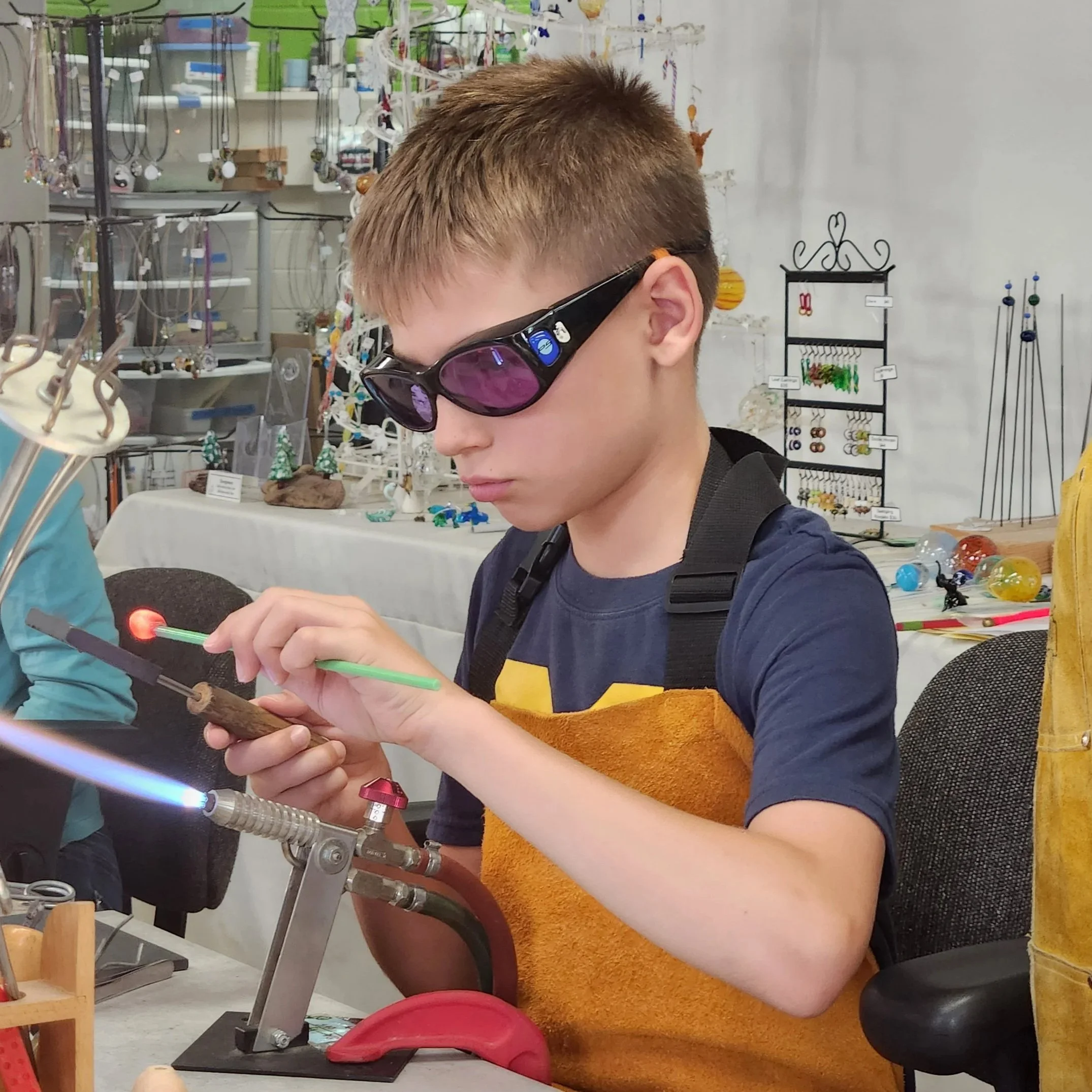 A young person with short brown hair and wearing black sunglasses is shopping for jewelry at a store. They are holding a green stick or tool while examining a ring on display. The background shows a variety of jewelry and decorative items on shelves and stands.