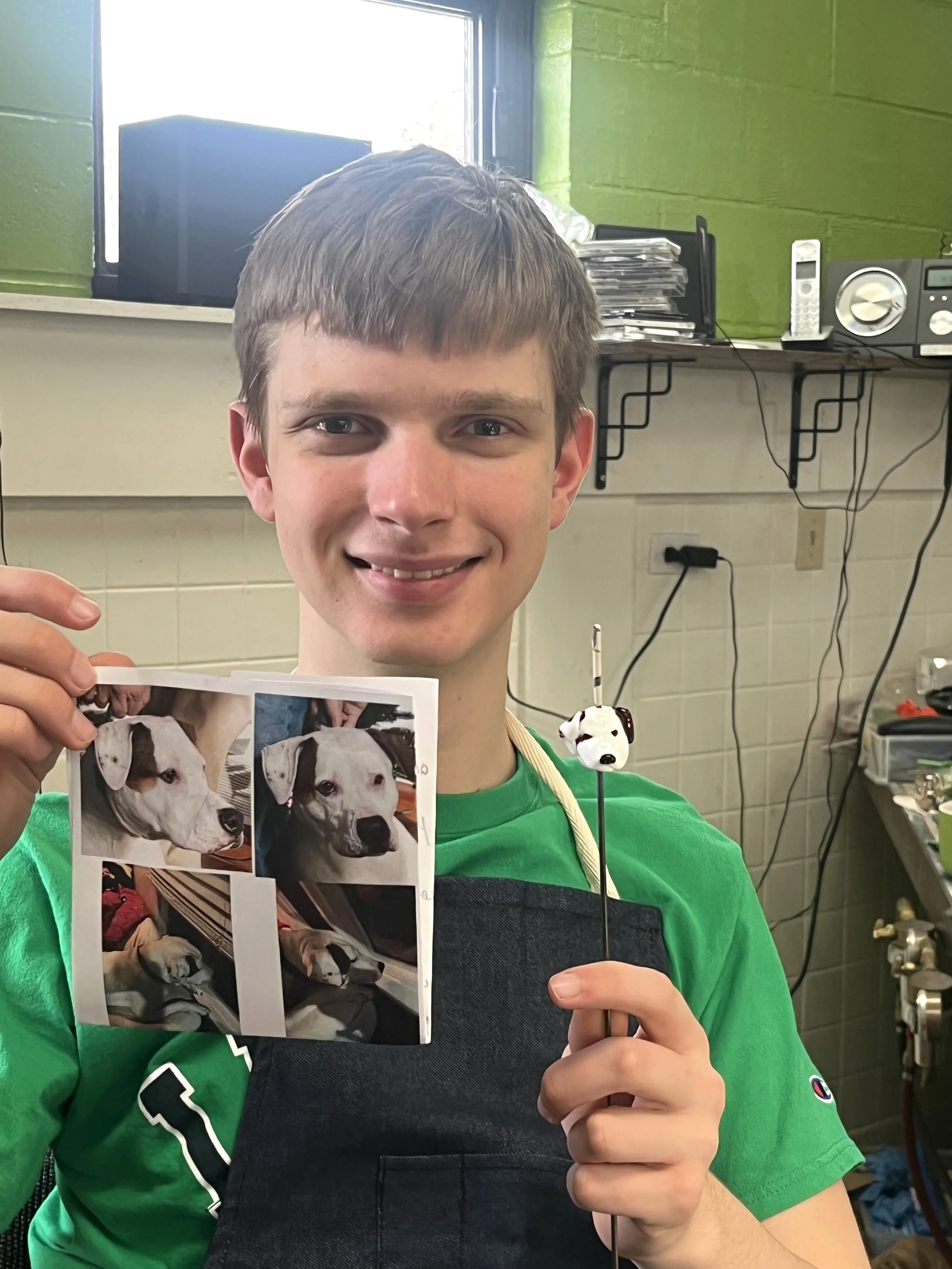 A young person with short brown hair and a big smile is holding a photograph of a dog and a small sculpture of a dog head. They are in a room with a green wall and a window, and there are various objects like a speaker, a telephone, and some papers in the background.