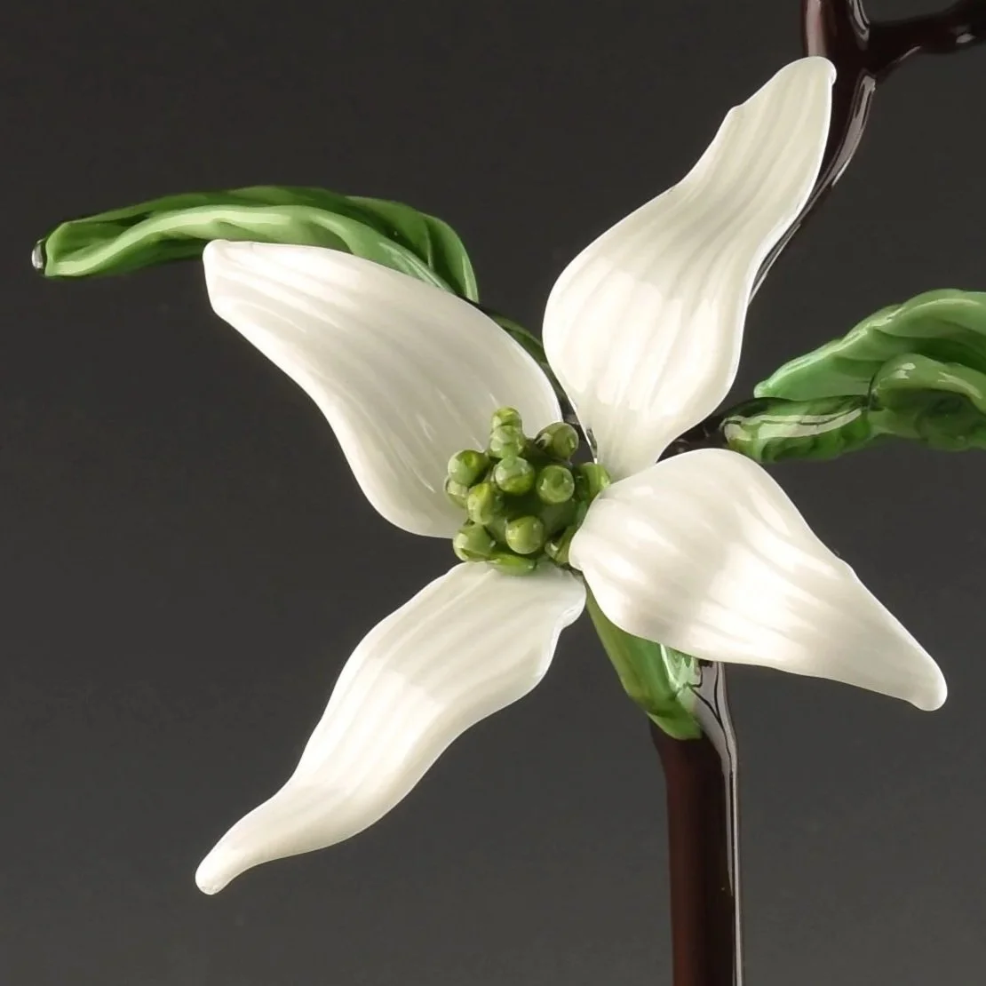 A close-up of a decorative piece resembling a white flower with six elongated petals, green leaves, and a dark stem, with dark liquid being poured onto it.