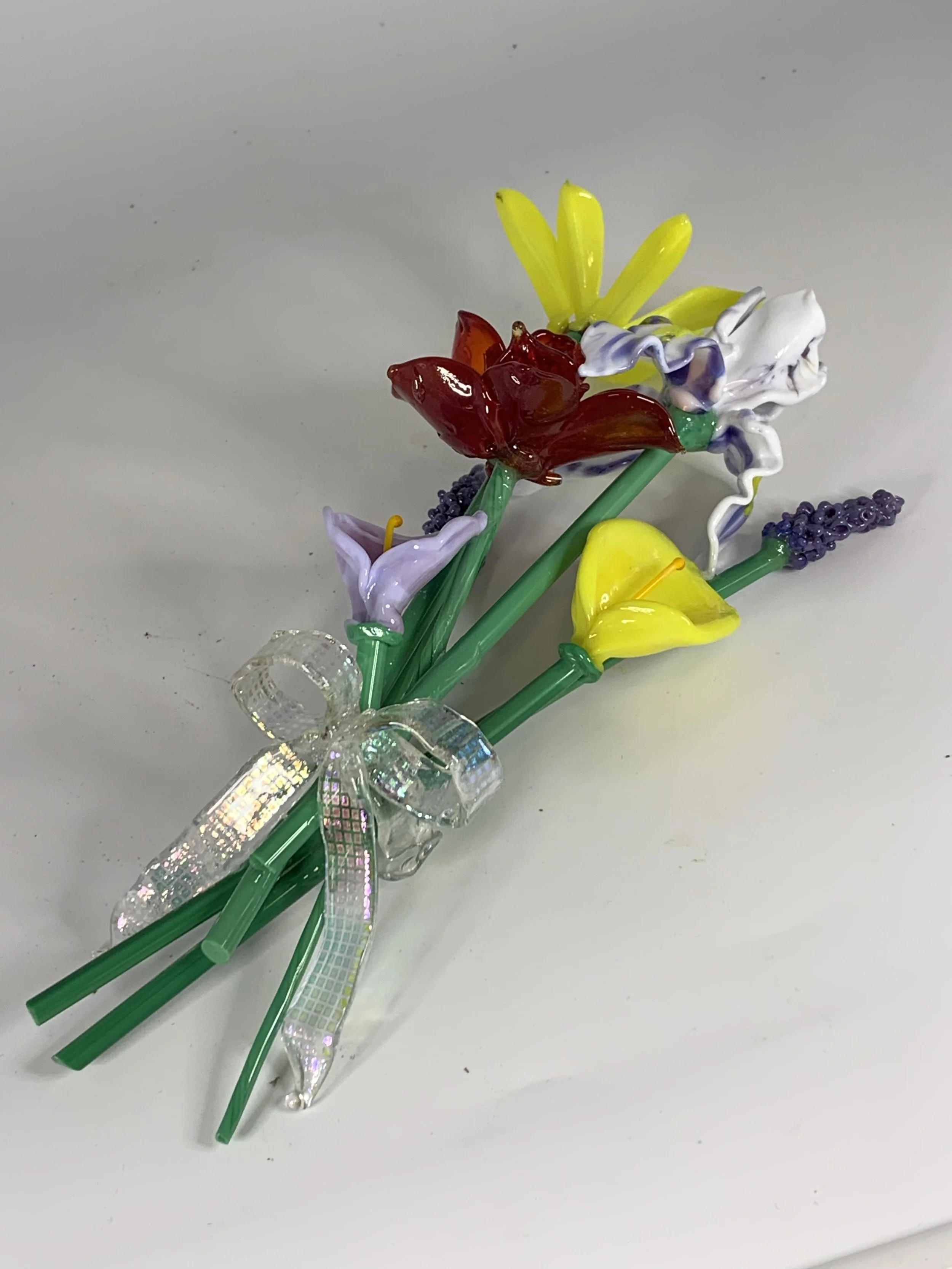 Colorful glass flowers arranged with green stems and a decorative bow on a white surface.
