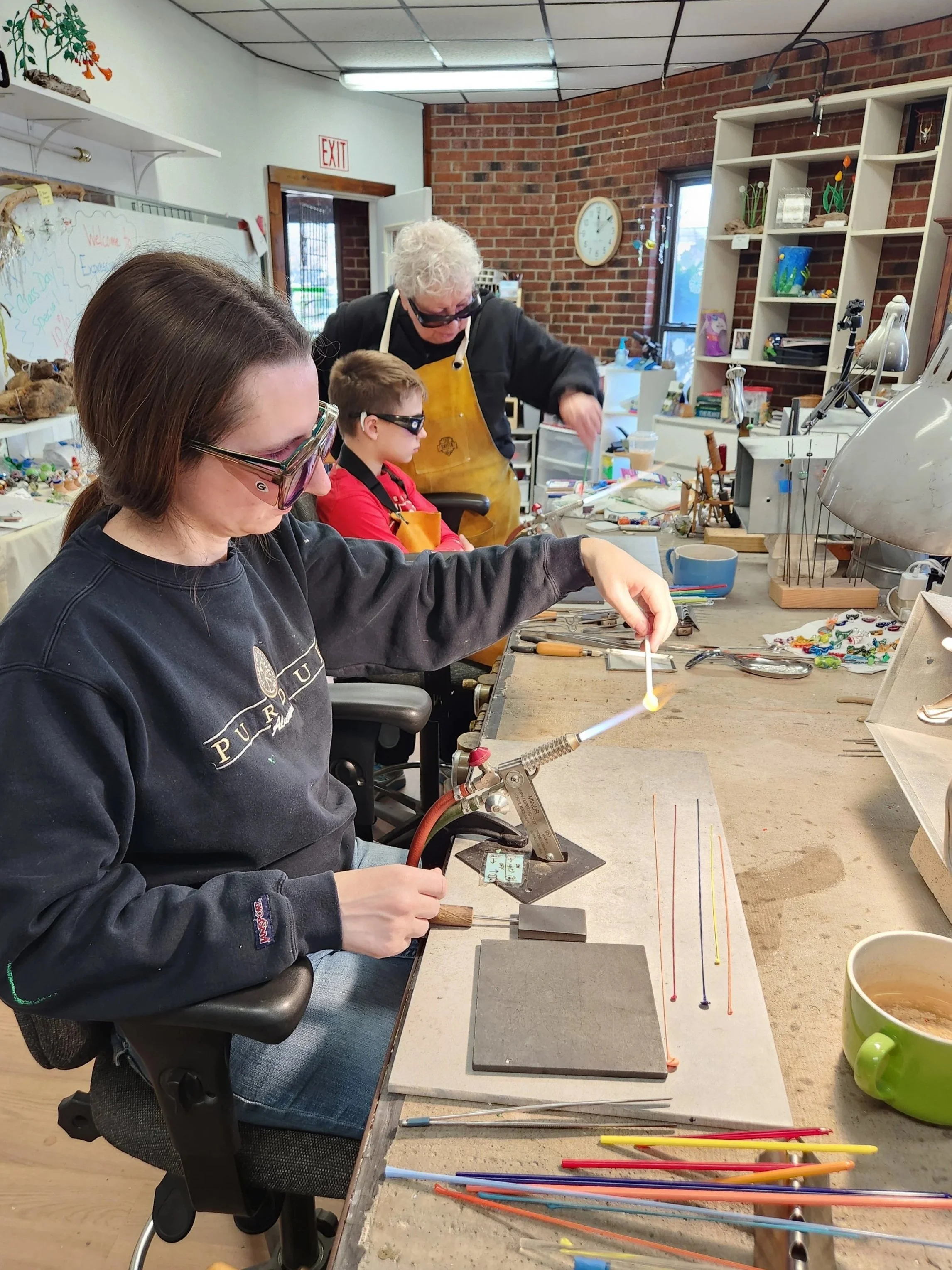 A woman and two children at a crafting table, working on glasswork with a flame, in a room with brick walls and shelves filled with art supplies and decorations.