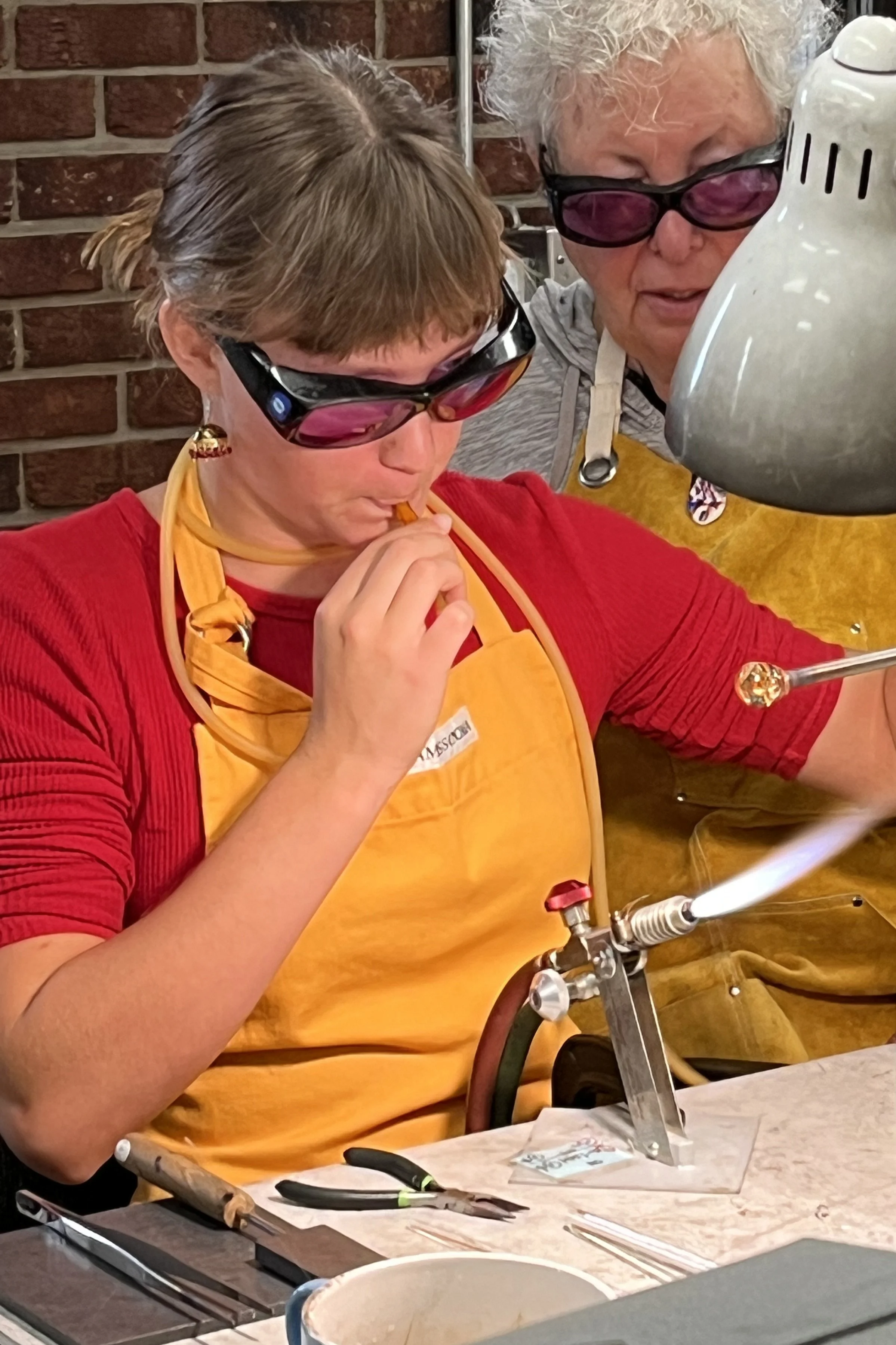 Two women wearing safety glasses working carefully with tools and equipment on a project at a workshop or art studio, with a brick wall in the background.