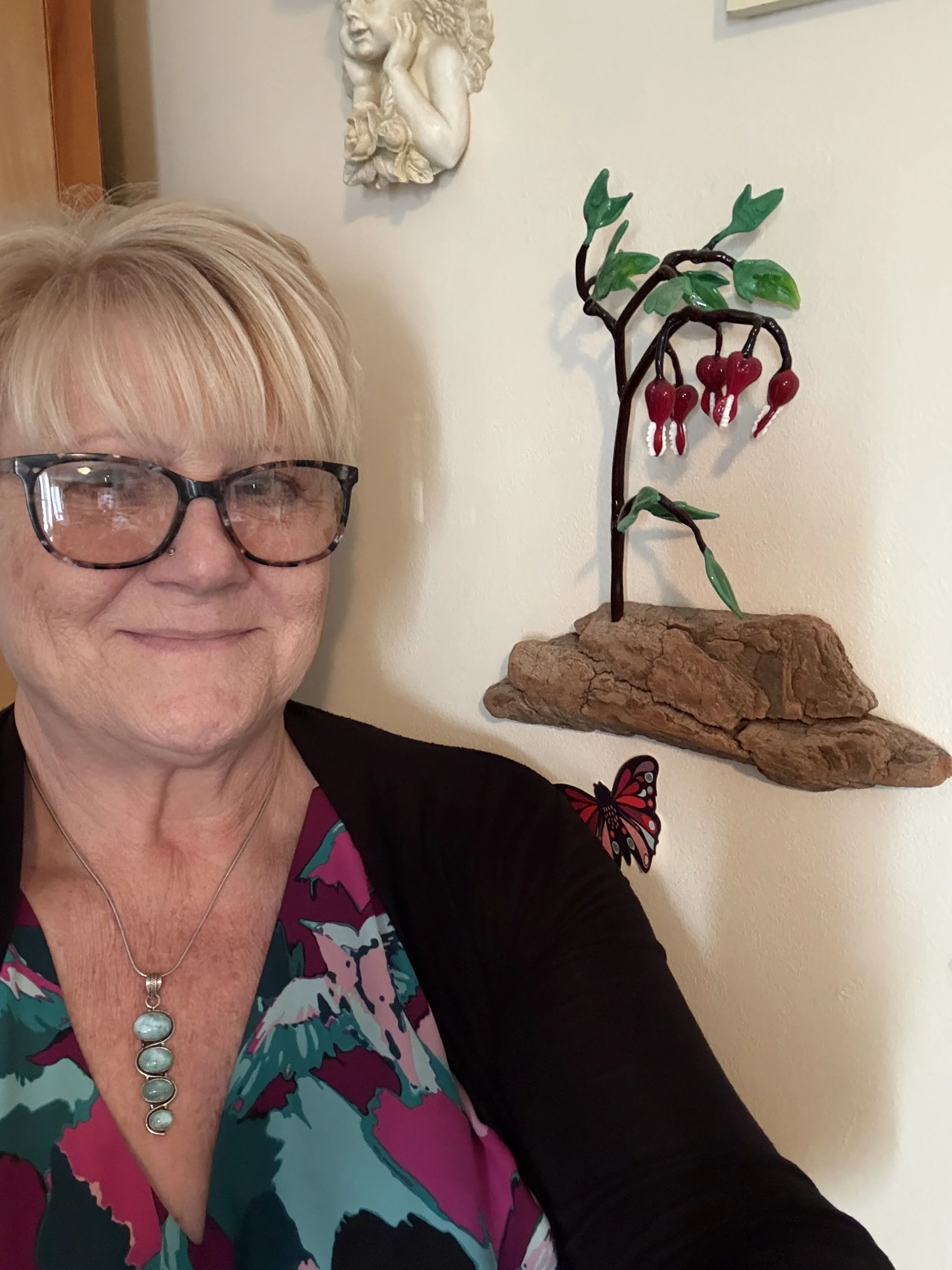 A woman with short blonde hair, glasses, and a floral blouse with pink, teal, and black colors, wearing a silver necklace with large oval stones, standing next to a metal sculpture of a cherry tree with green leaves and red cherries, mounted on a rock on the wall.