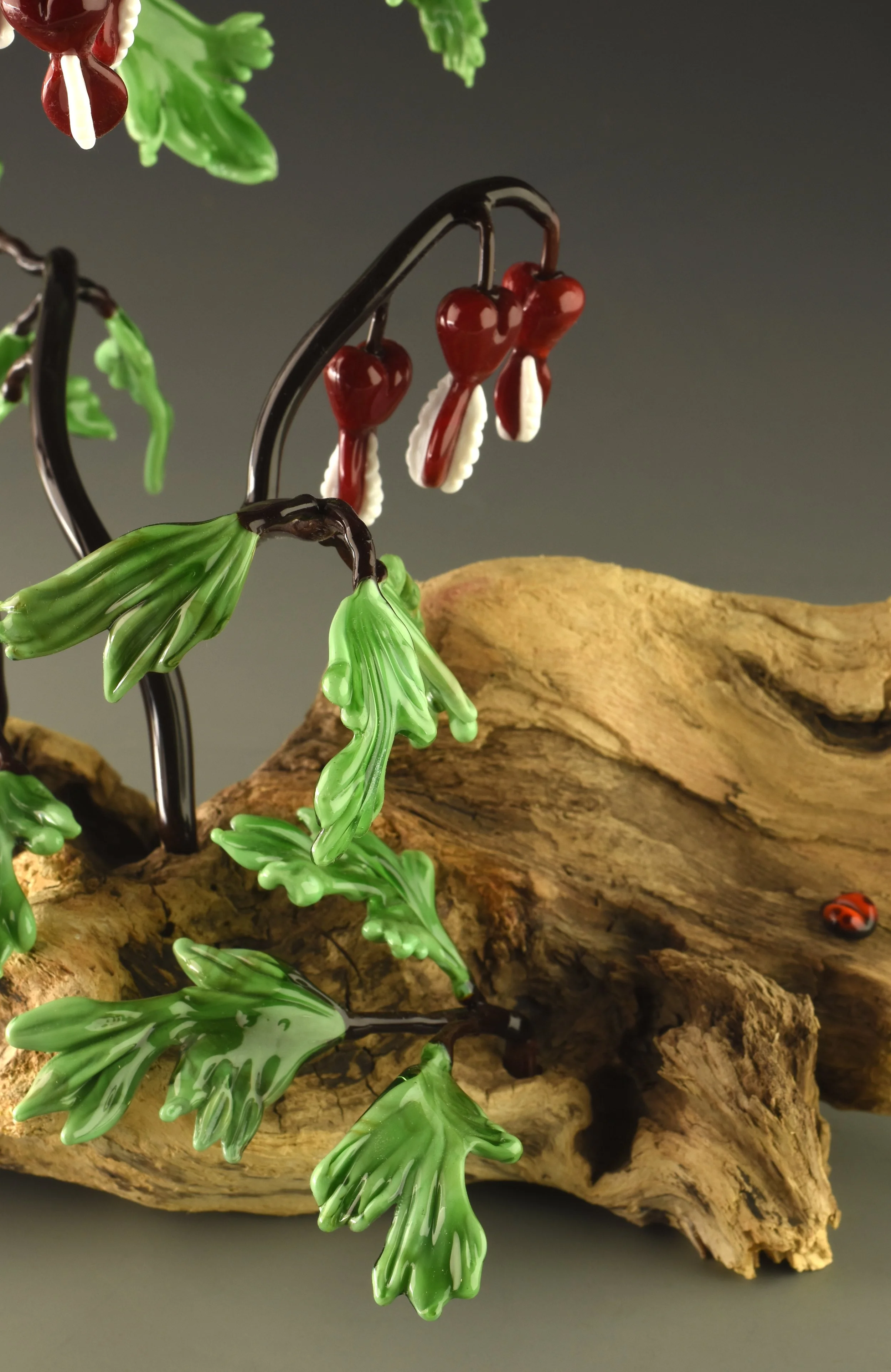 A decorative glass art piece resembling a plant with green leaves, red hearts, and white accents, mounted on a piece of textured wood.