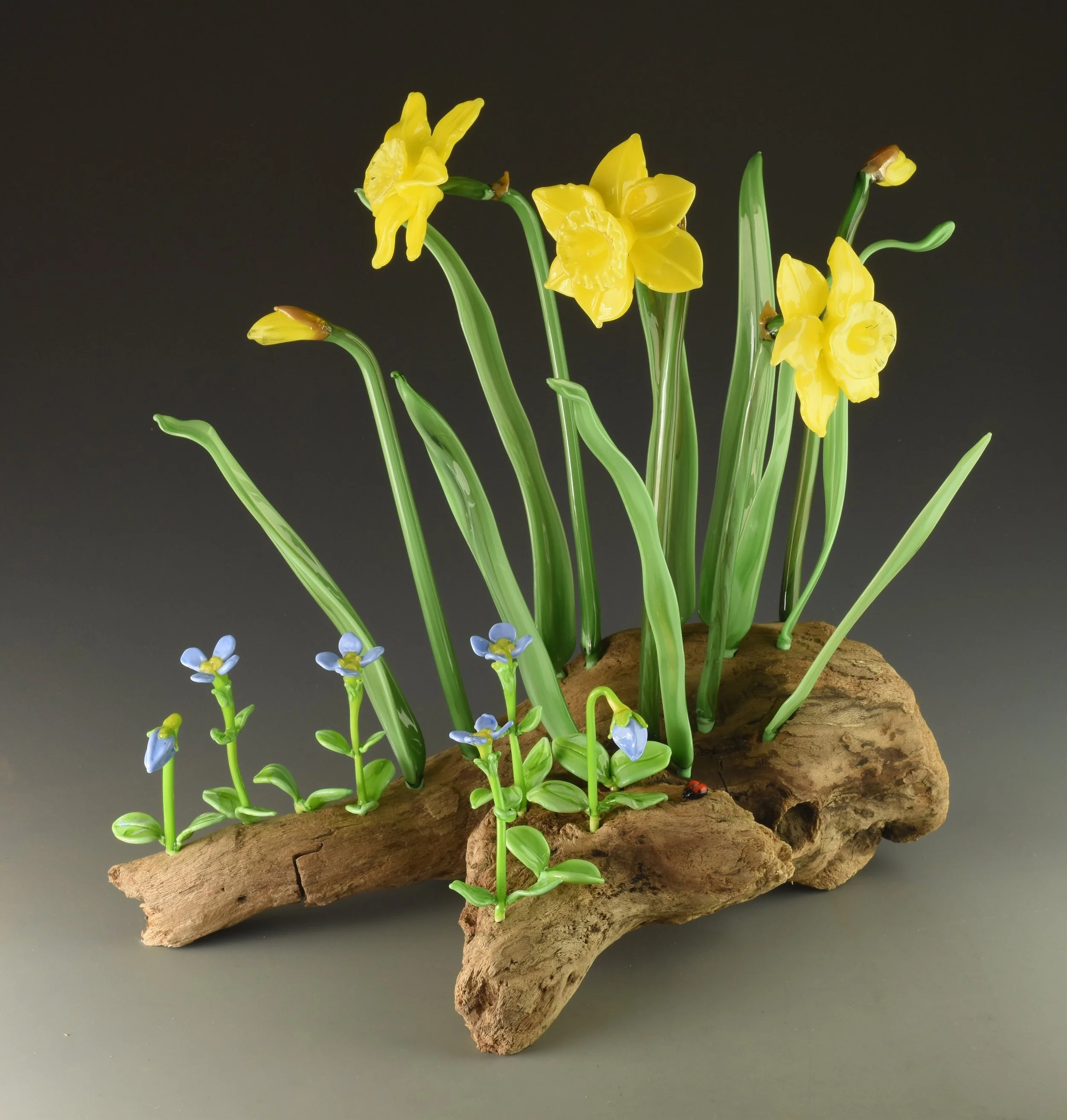 Glass sculpture of yellow daffodil flowers with green stems and leaves, mounted on a piece of natural wood.