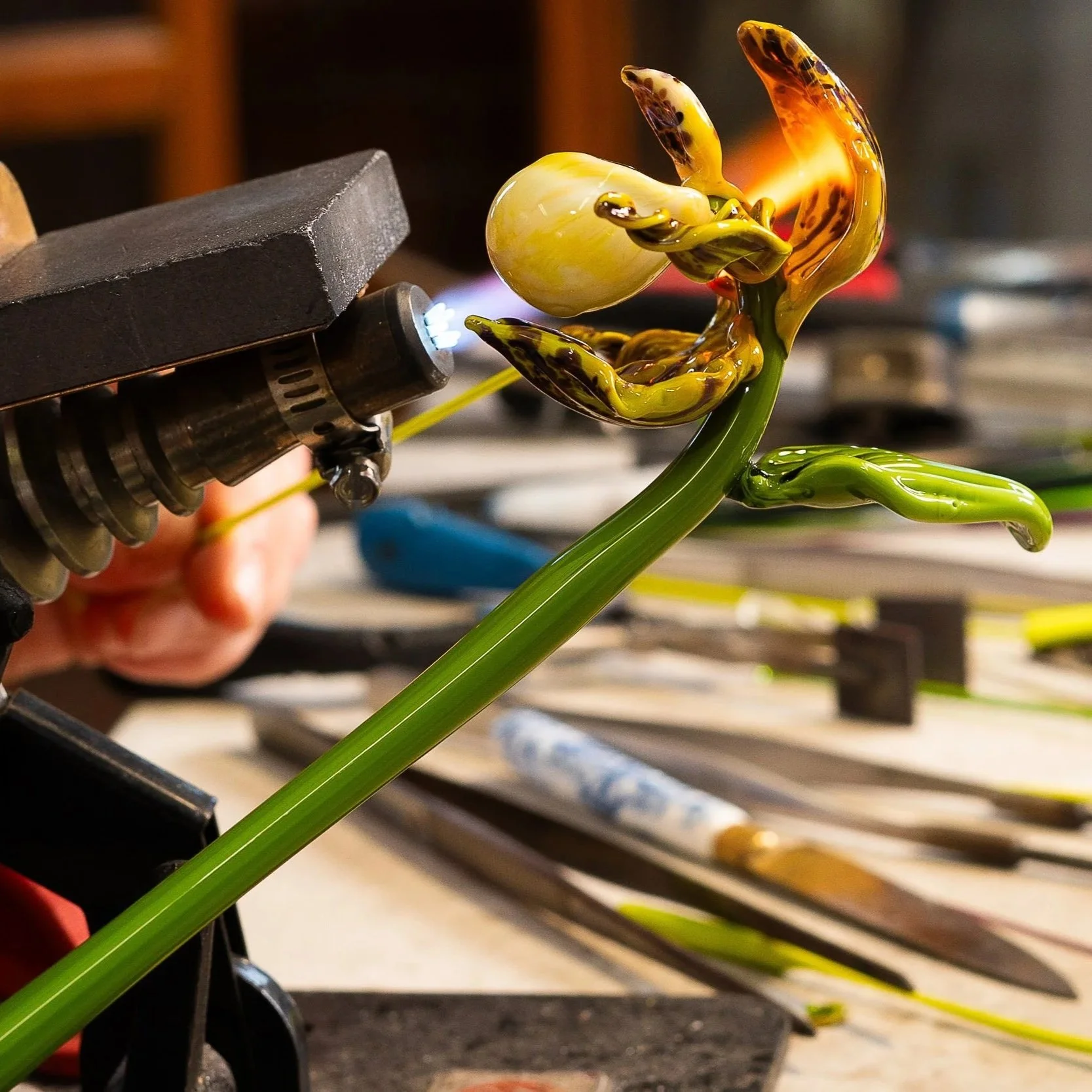 A glassblowing process with a glowing orange and yellow molten glass shape being formed on a green glass stem, with tools and equipment in the background.