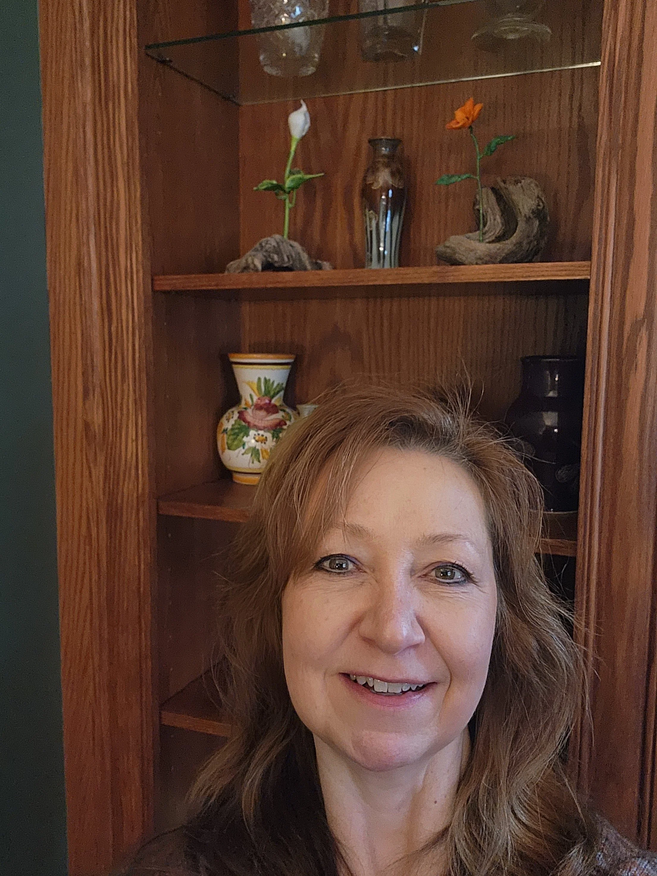 A woman with light skin and curly light brown hair taking a selfie in front of a wooden display cabinet. The top shelves of the cabinet contain decorative glassware and vases, including two small vases with flowers. The woman is smiling and looking at the camera.