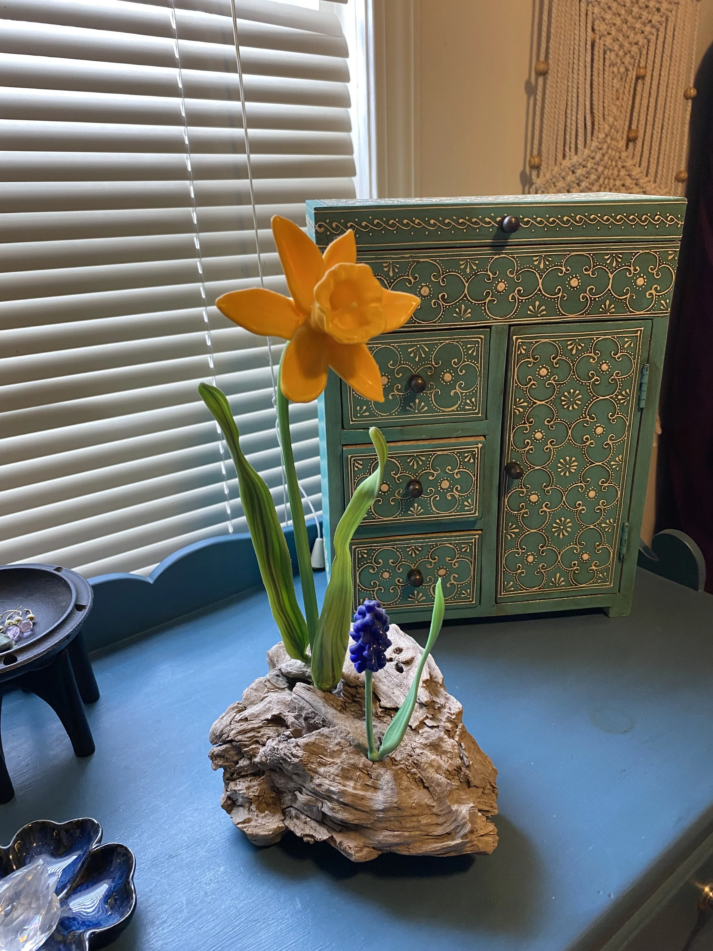Yellow daffodil and purple hyacinth flowers on a rock base in front of a decorative cabinet, with blinds and a macrame wall hanging in the background.