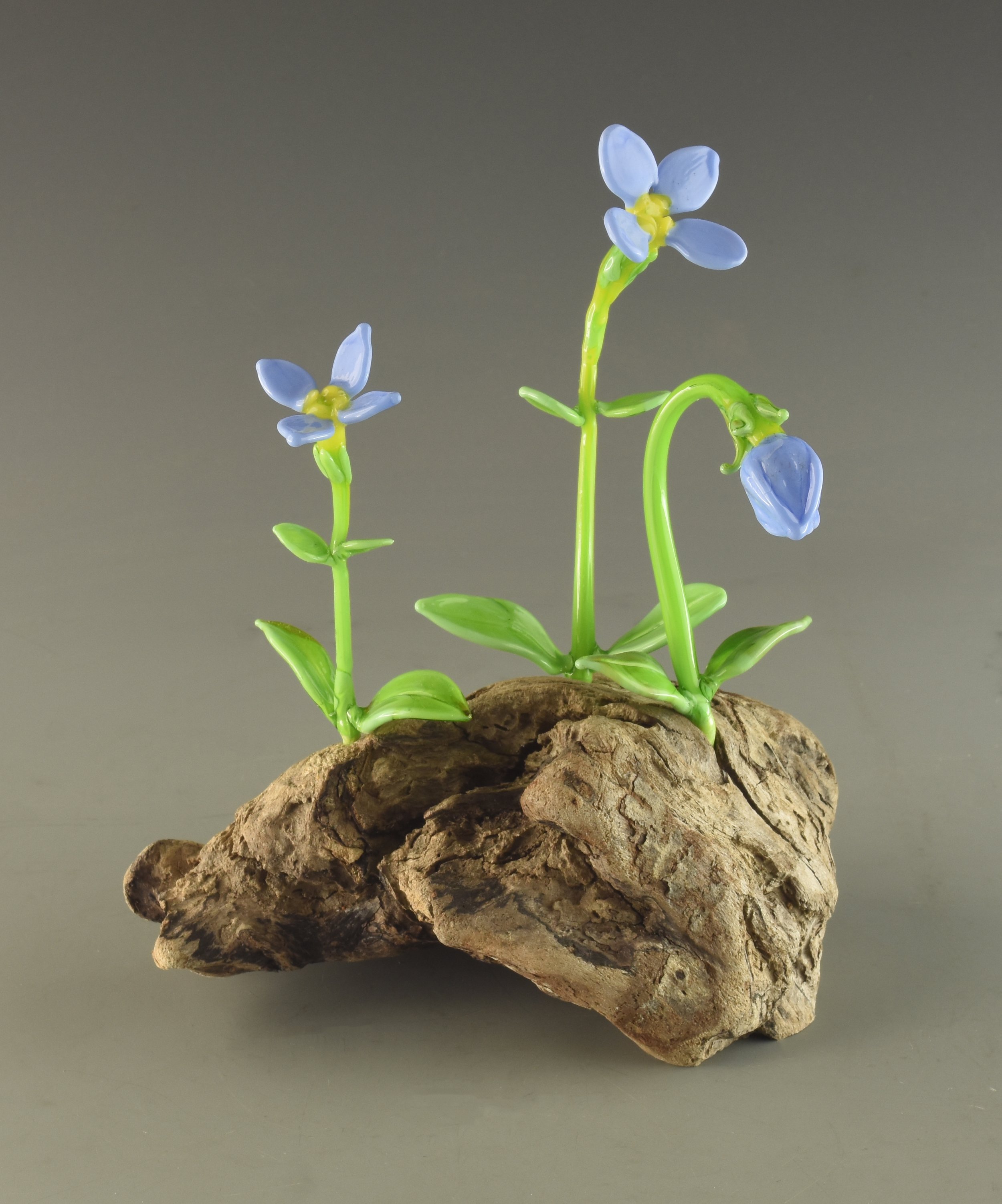 A small arrangement of blue artificial flowers with green stems and leaves, attached to a piece of rough, natural wood, set against a plain gray background.