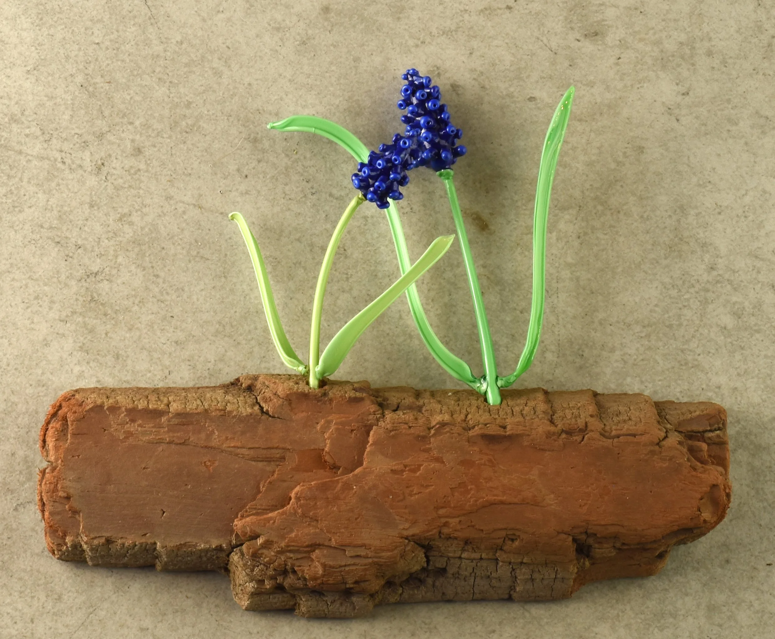 A handcrafted model of a flowering plant made from twisted green and blue plastic pieces attached to a piece of wood.