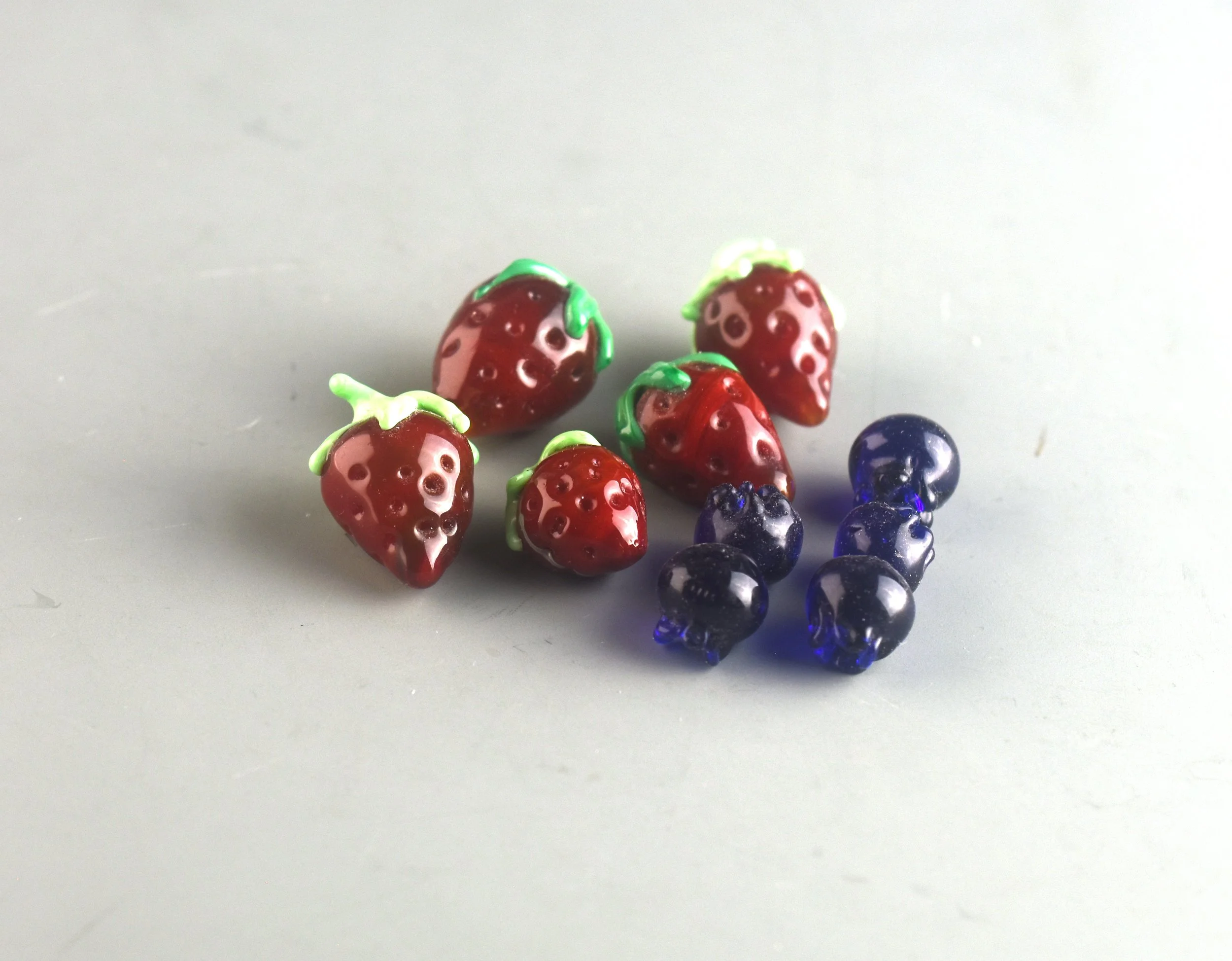 Close-up of small glass strawberry and blueberry figurines on a plain surface.