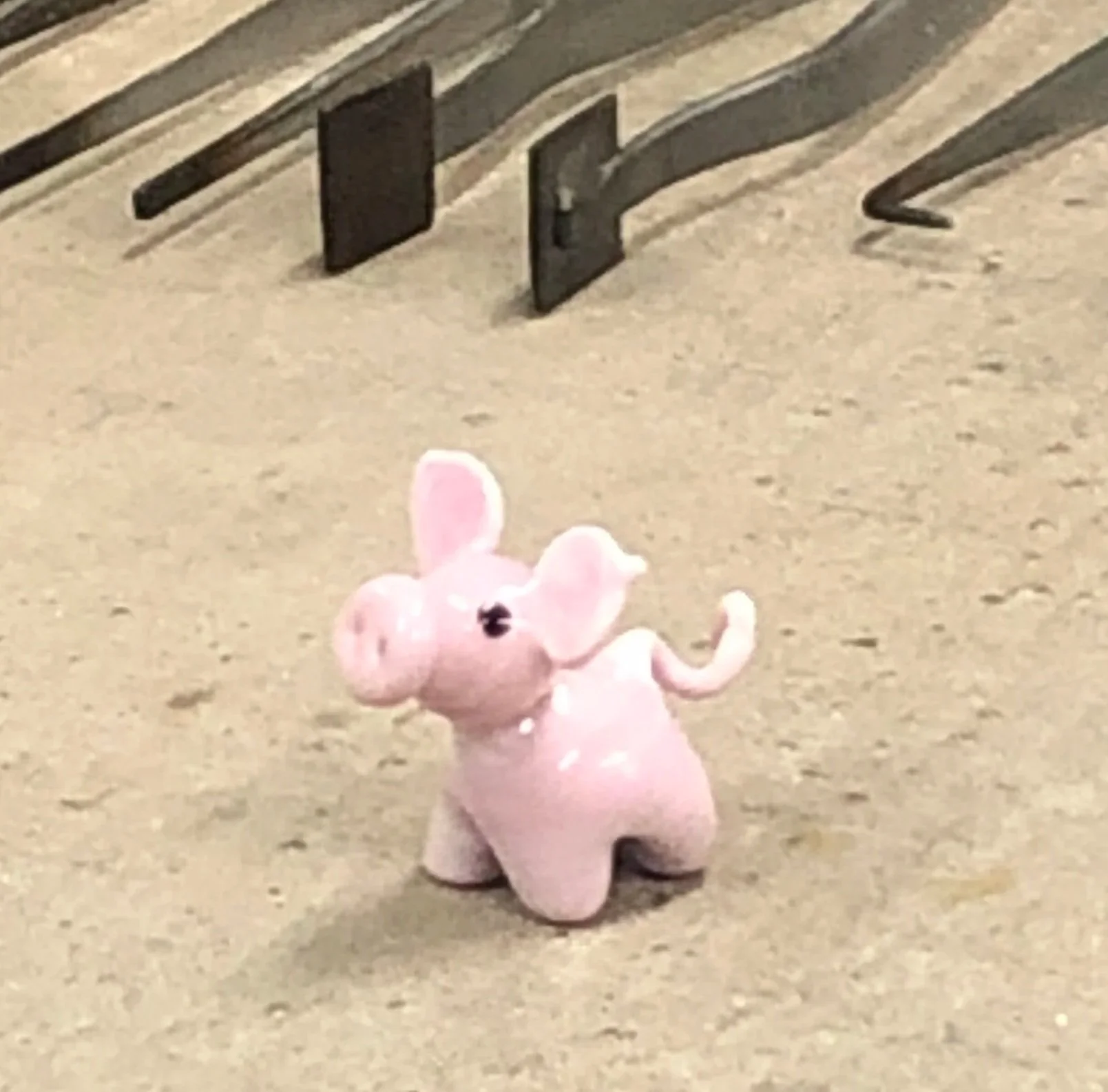 Small pink toy elephant with large ears and black eyes standing on a sandy surface, with a black rectangular object and a lock visible in the background.