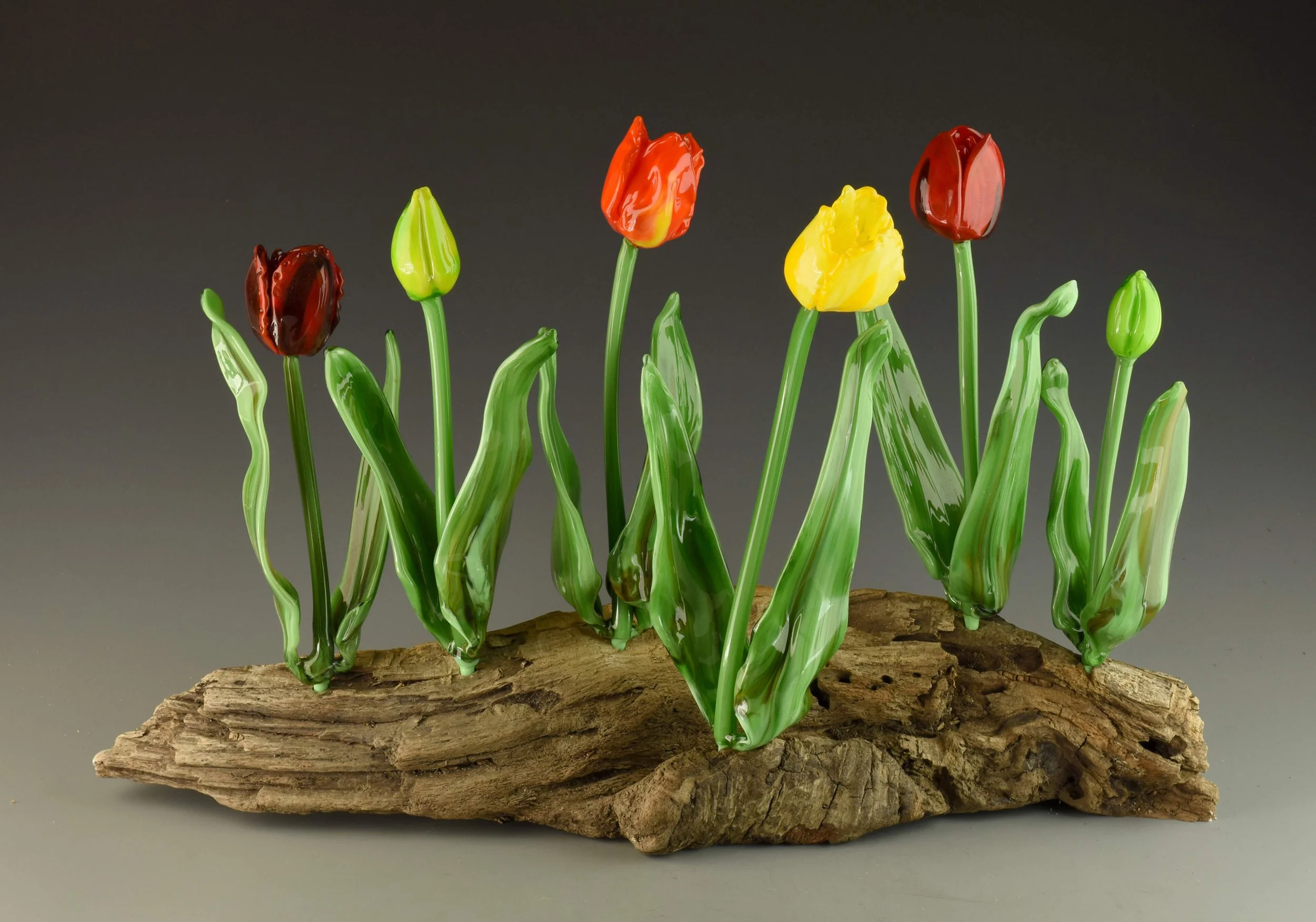 Glass art tulips with green stems on a piece of wood, set against a dark background.