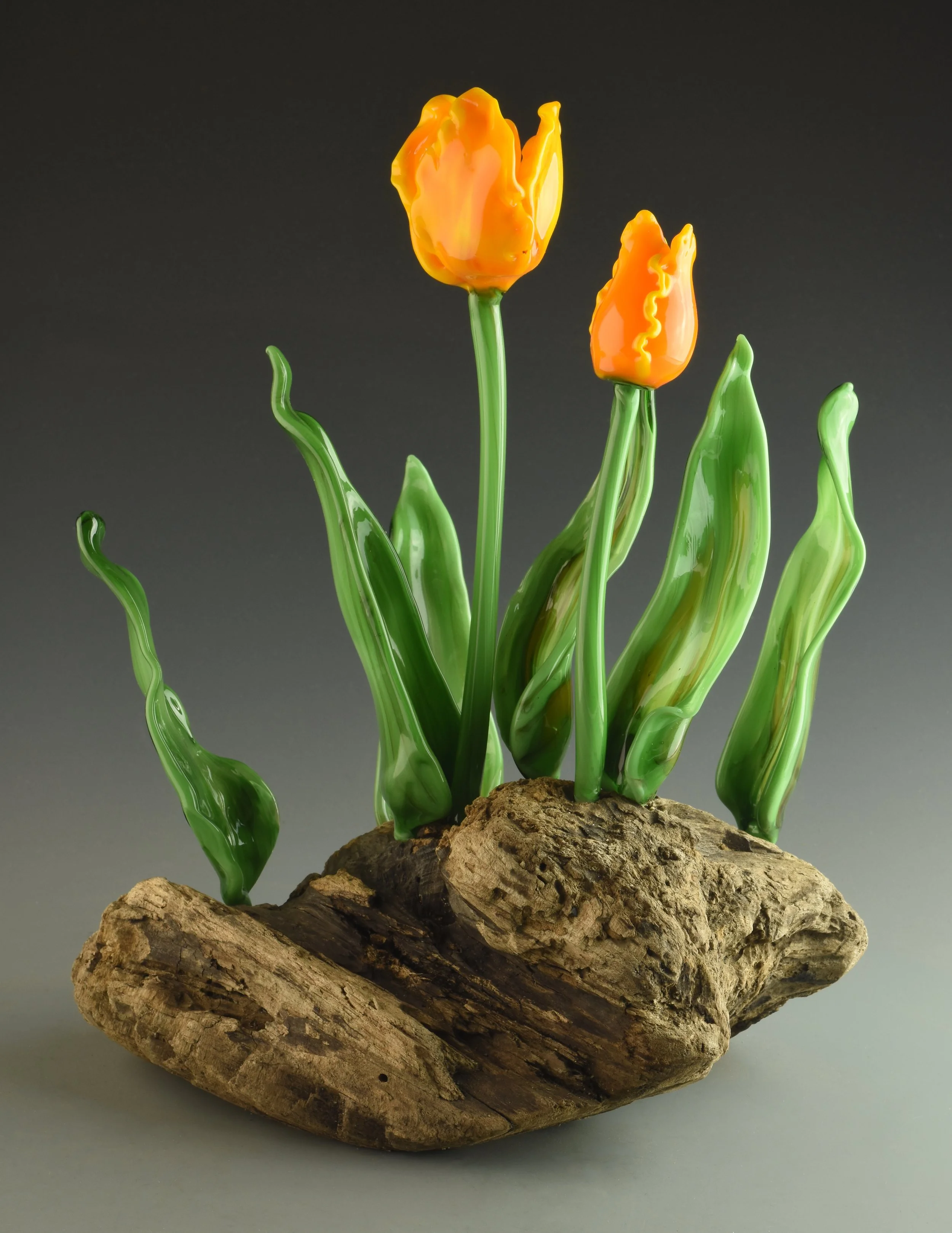 Glass tulip flowers with green stems and leaves on a piece of wood against a gray background.