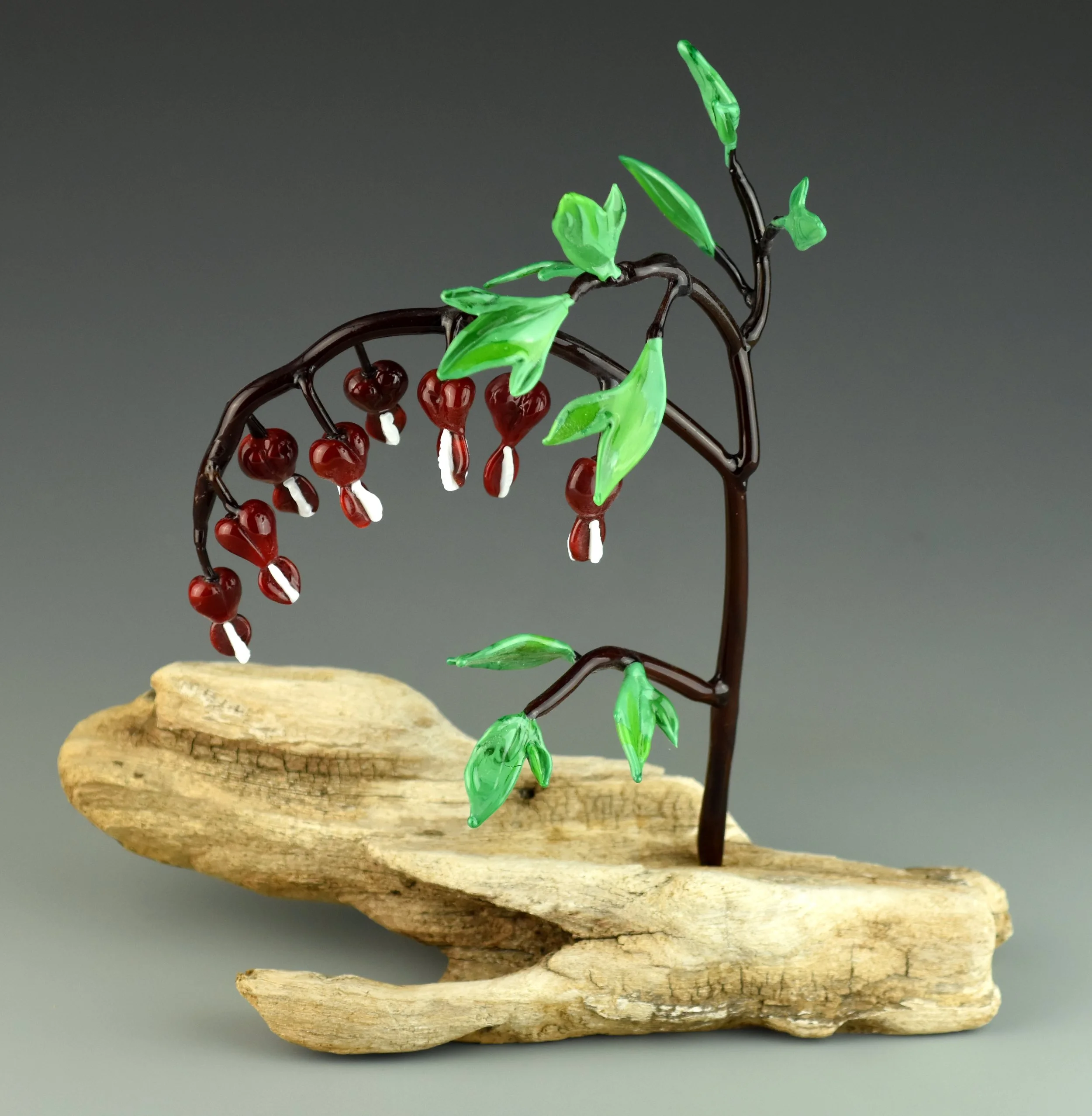Glass sculpture of a bird perched on a branch with green leaves, mounted on a piece of driftwood.