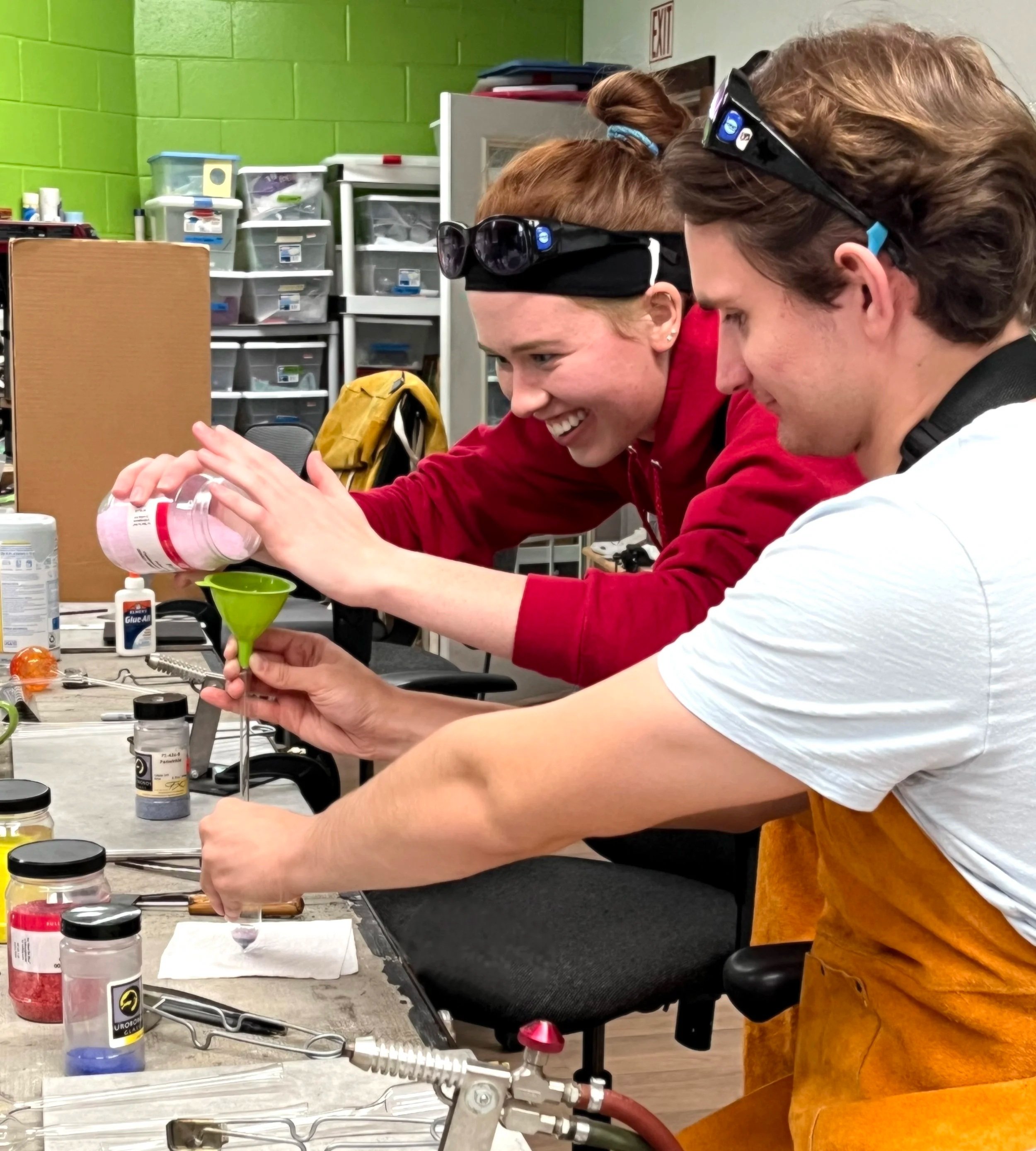 Two glass art students smiling while working in a glass art studio with tools and supplies on the worktable.