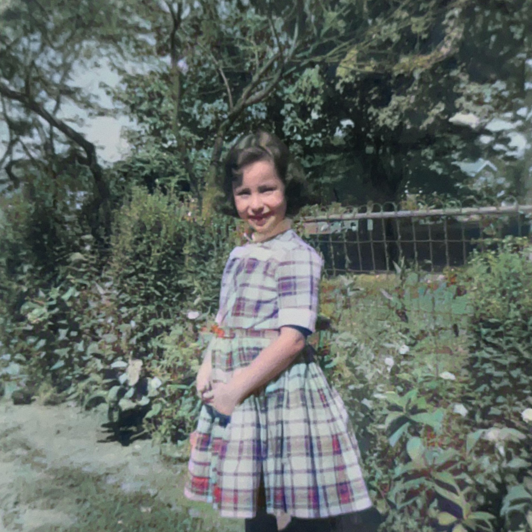 A young girl with dark wavy hair smiling in a garden, wearing a vintage plaid dress with short sleeves, standing near a low hedge and a wooden fence, surrounded by greenery and trees in the background.