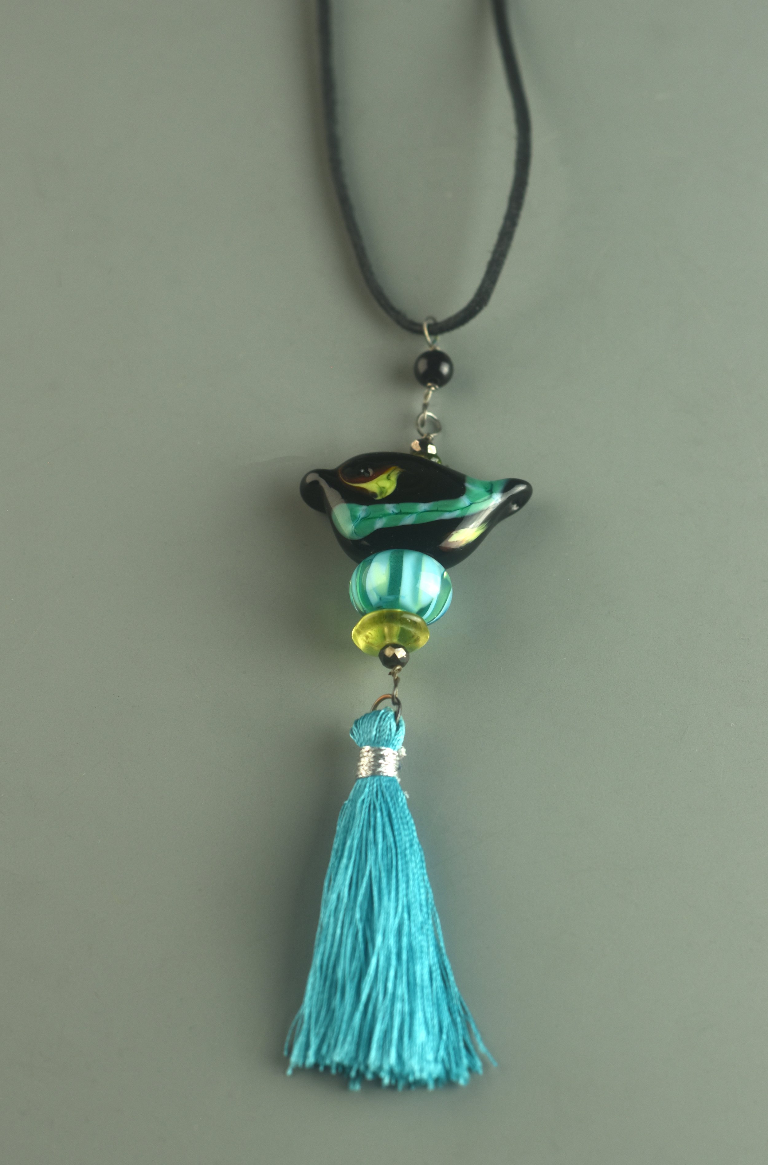 Colorful beaded and tassel wall hanging with a black and green glass bead, a glass figure resembling a bird or fish, and a large turquoise tassel.