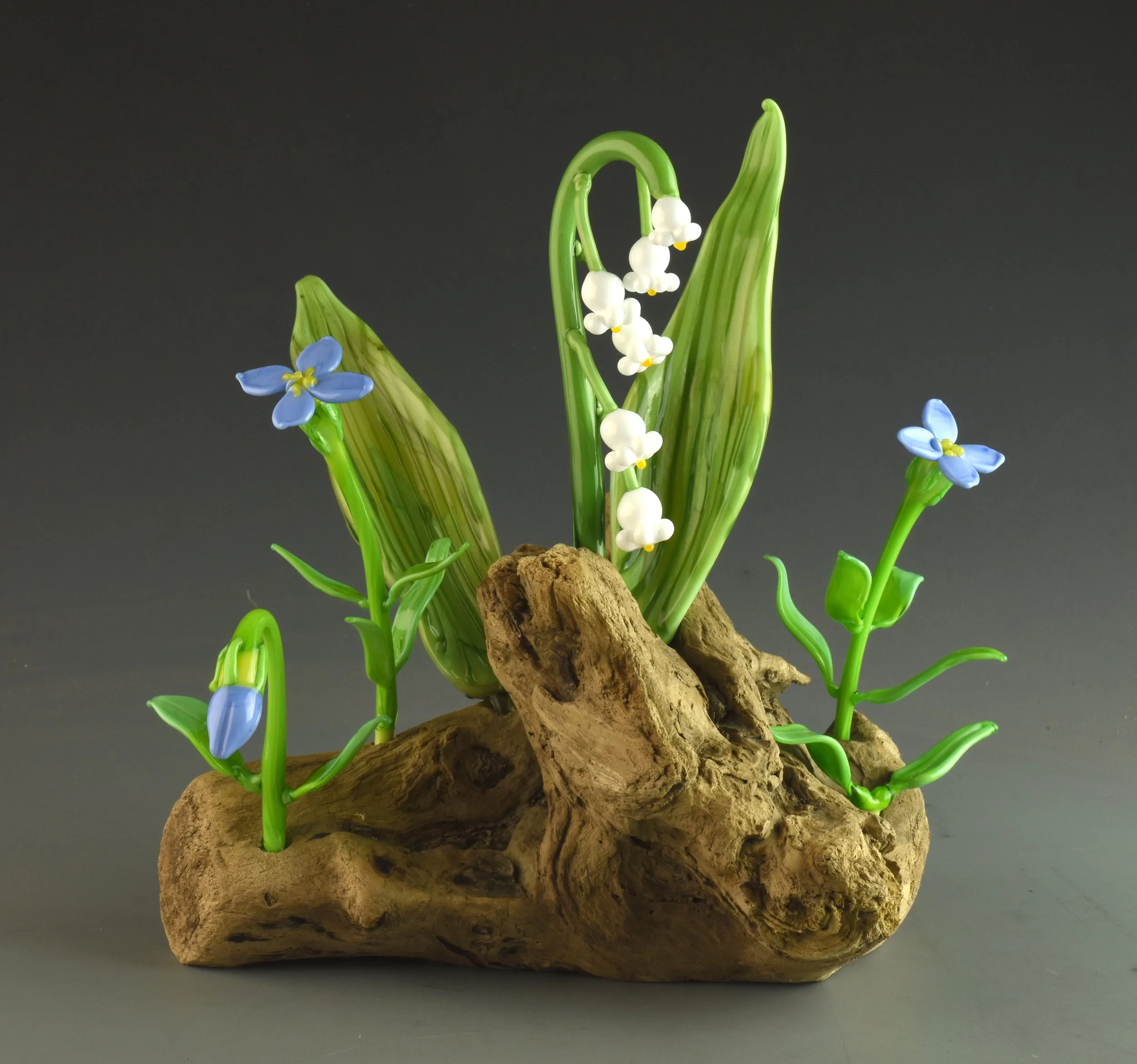 Glass decorative flowers resembling lily of the valley and blue forget-me-nots, arranged among green leaves and mounted on a textured piece of wood.