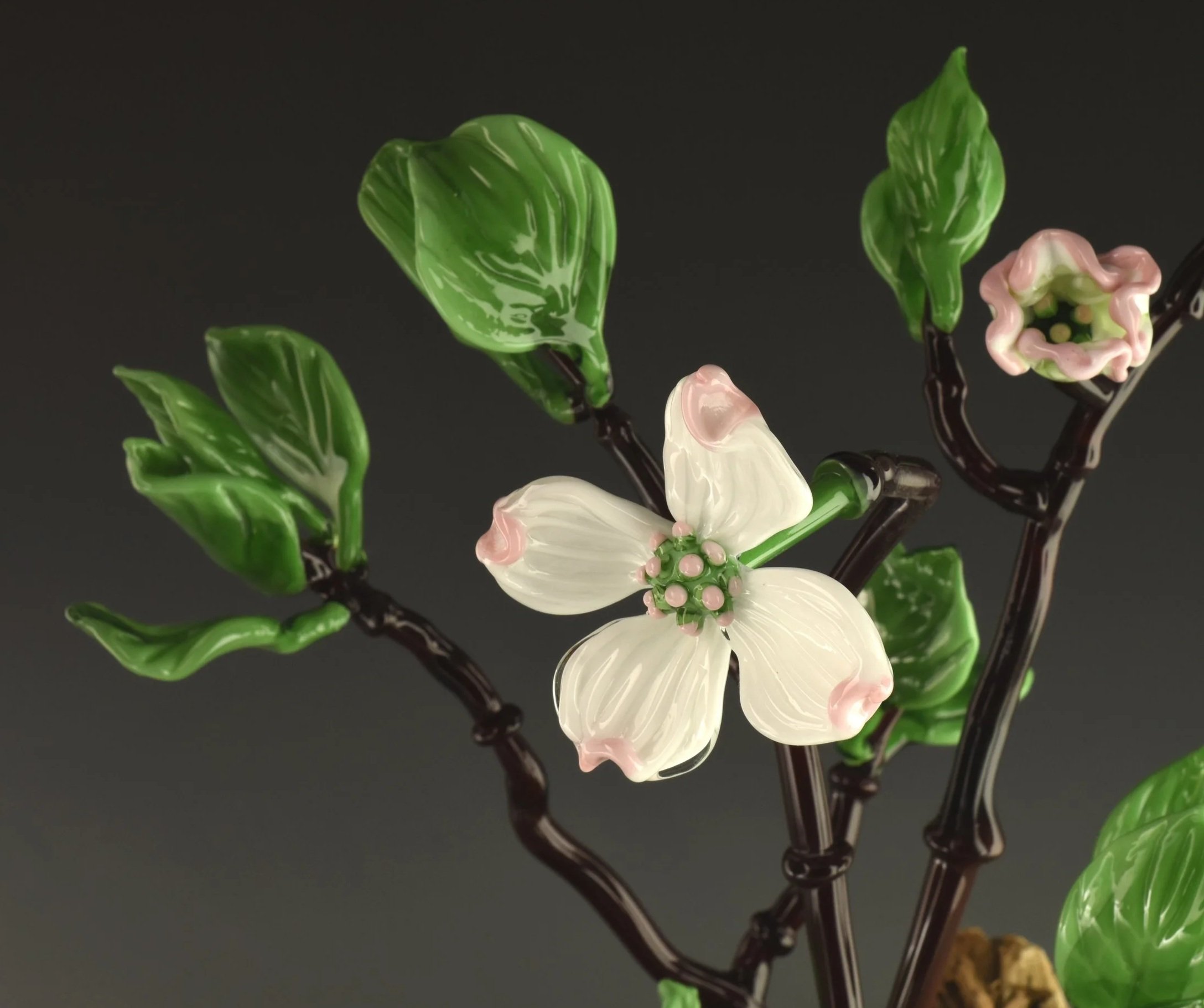 Close-up of decorative glass flowers, including pink and white blossoms with green leaves and dark stems, on a dark background.