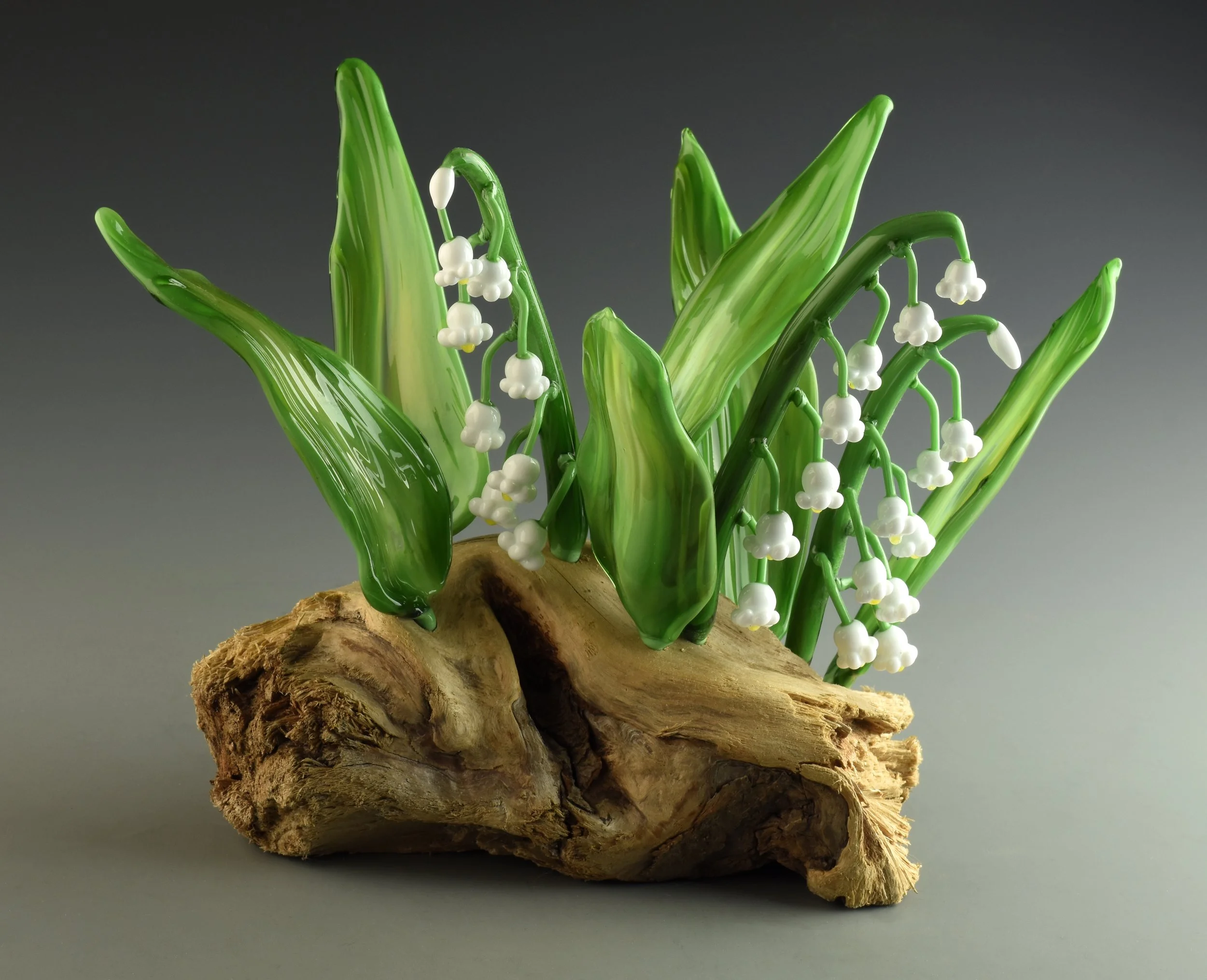 A decorative display of artificial lily of the valley flowers with green leaves and white bell-shaped blossoms, arranged on a piece of natural driftwood.