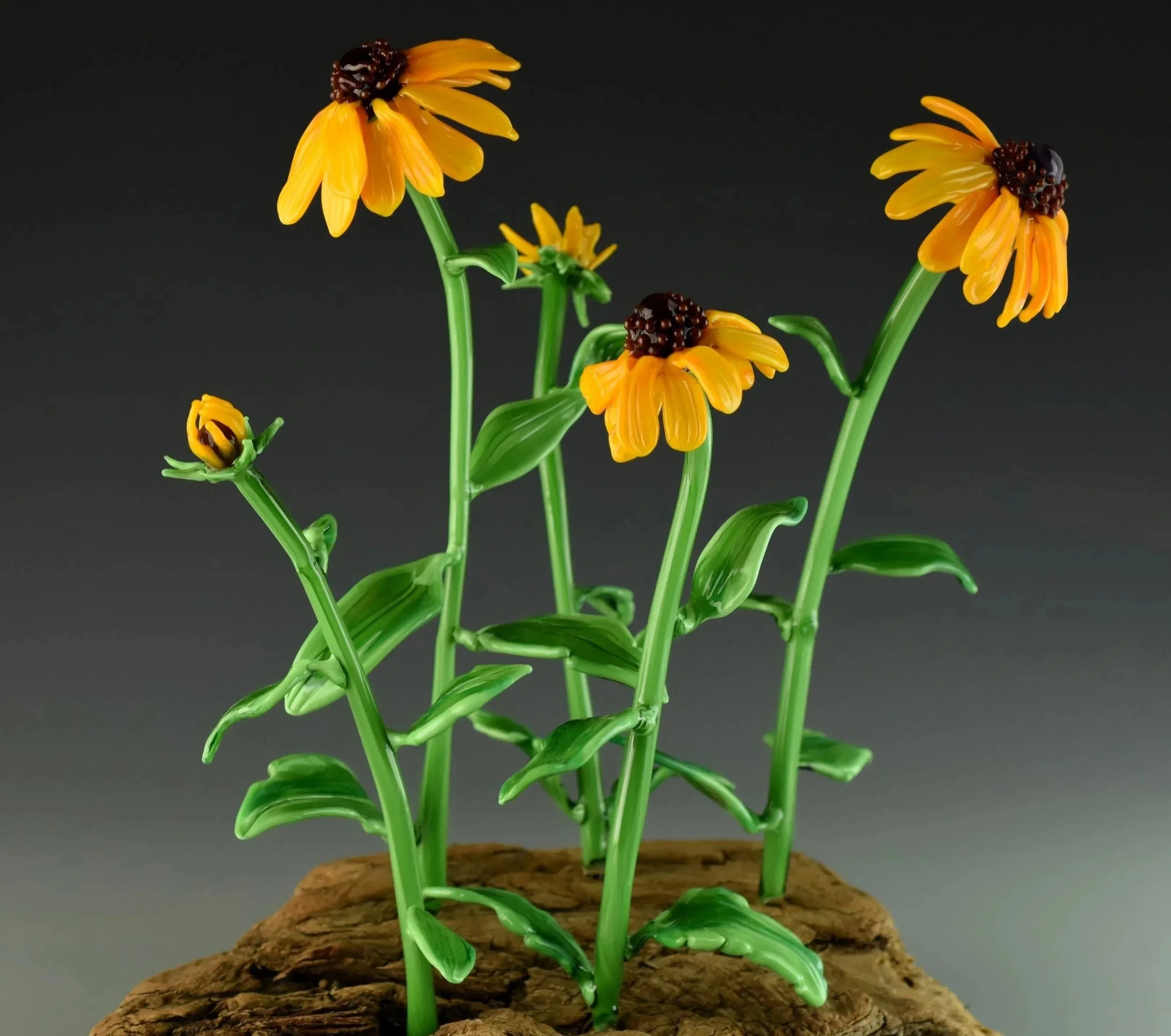 Glass sculpture of yellow flowers with dark centers on green stems, displayed on a textured brown base against a black background.