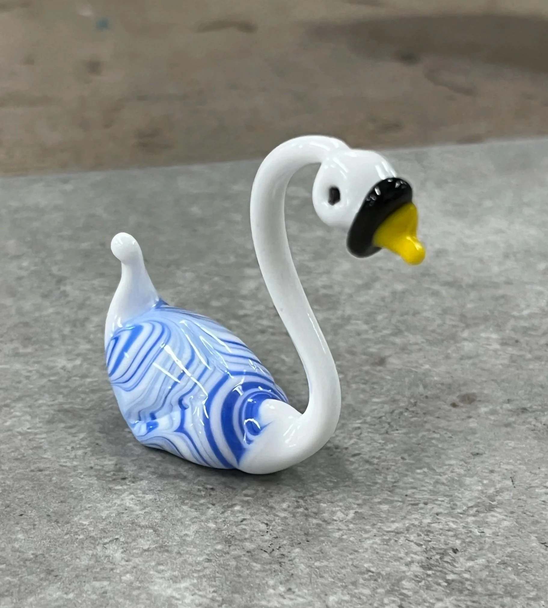 Glass swan figurine with a white body, blue and white marble pattern, and a long curved neck, standing on a gray surface.