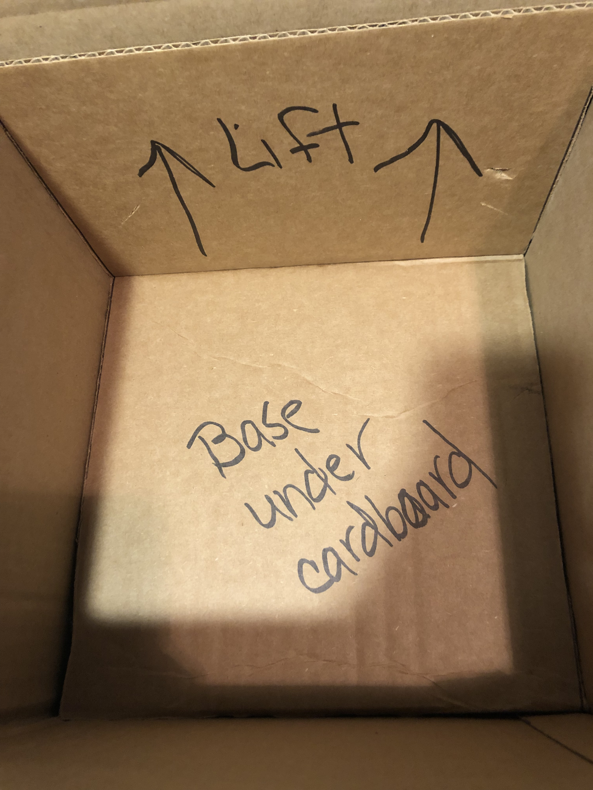Inside a cardboard box with handwritten words and arrows. The words 'Lift' with arrows pointing up and 'Base under' are visible.