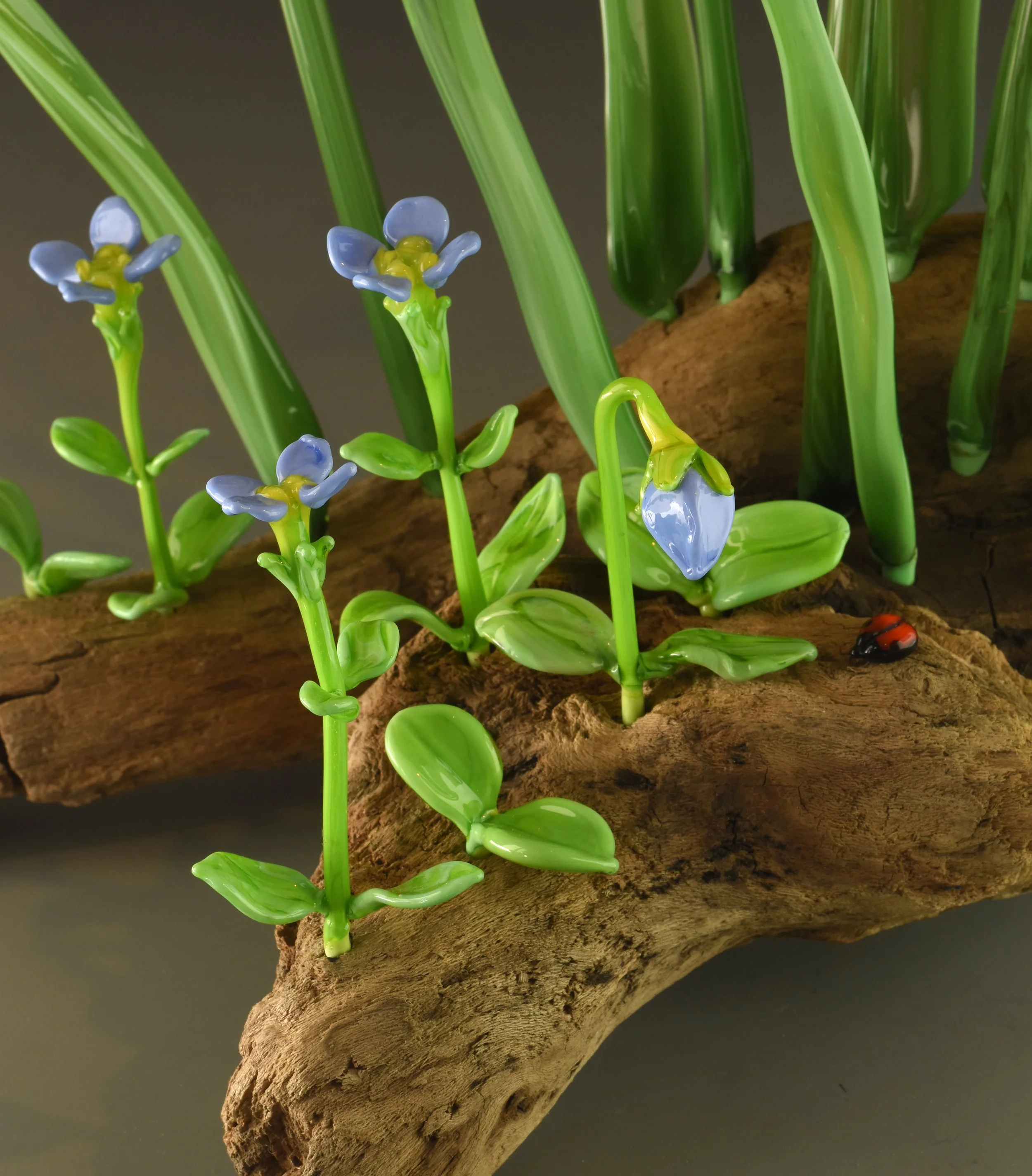 Close-up of a decorative glass plant with blue flowers and green leaves, mounted on a textured piece of driftwood.