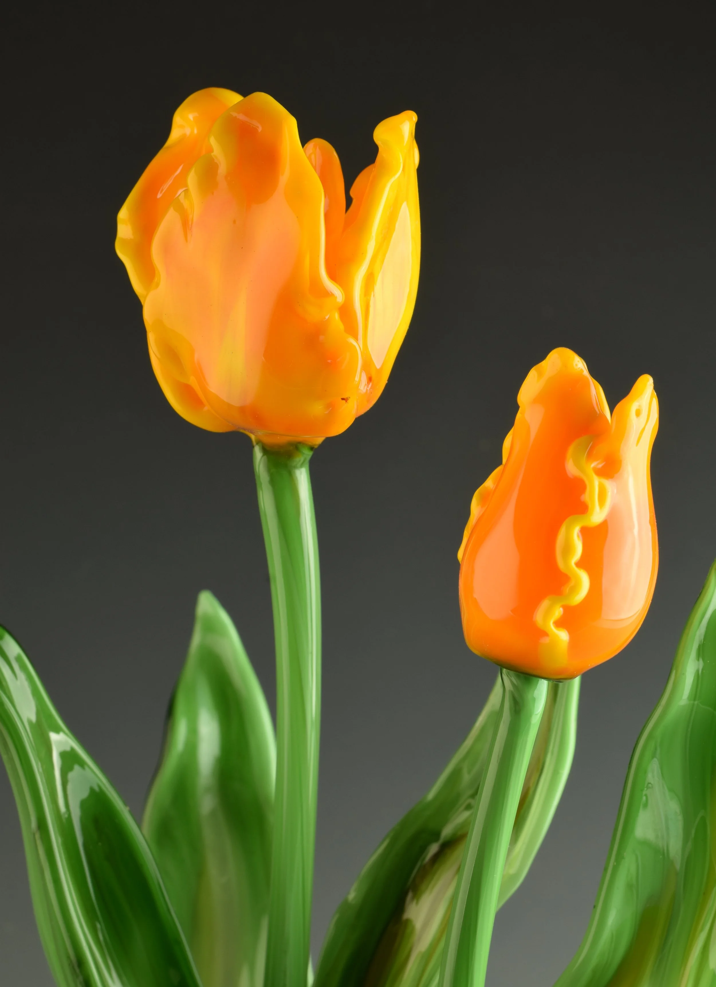 Close-up of three glass tulip flowers with orange and yellow petals and green stems on a dark background.