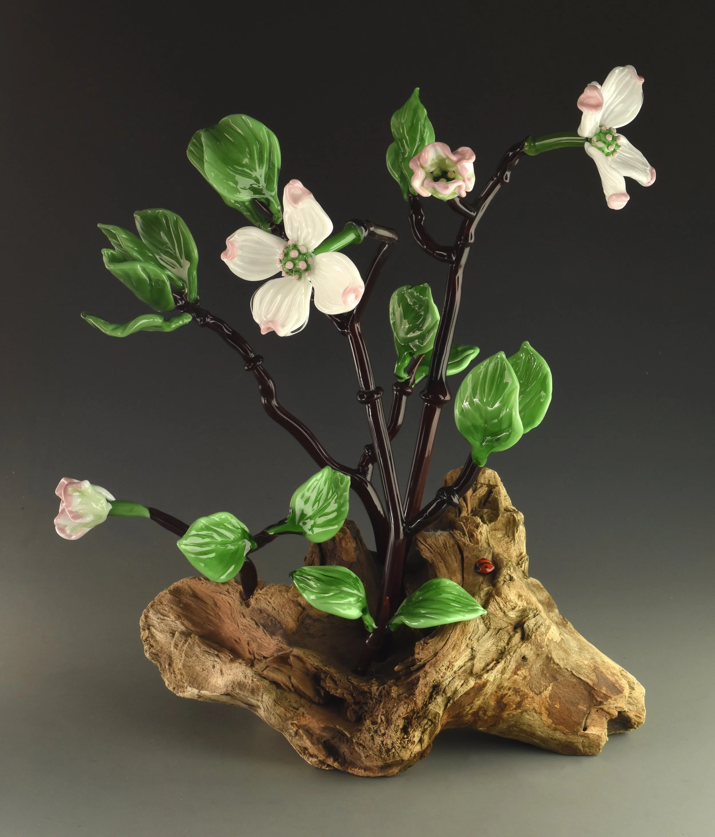 Decorative glass sculpture of flowering plant with white and pink flowers, green leaves, and dark stems mounted on a rough wooden base.