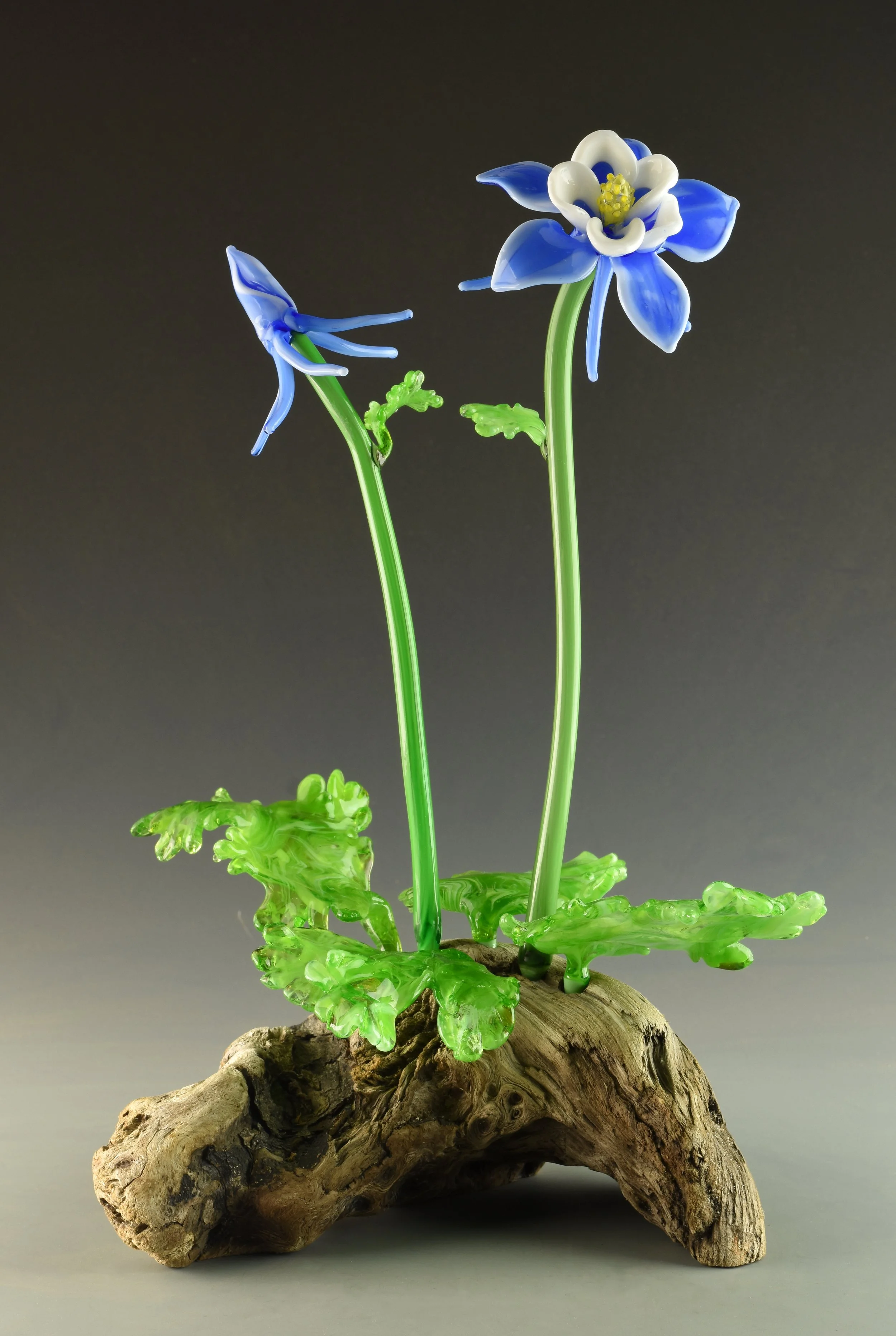 Glass art resembling a blue and white flower with green stems, set on a piece of textured wood with green glass leaves at the base, against a dark background.